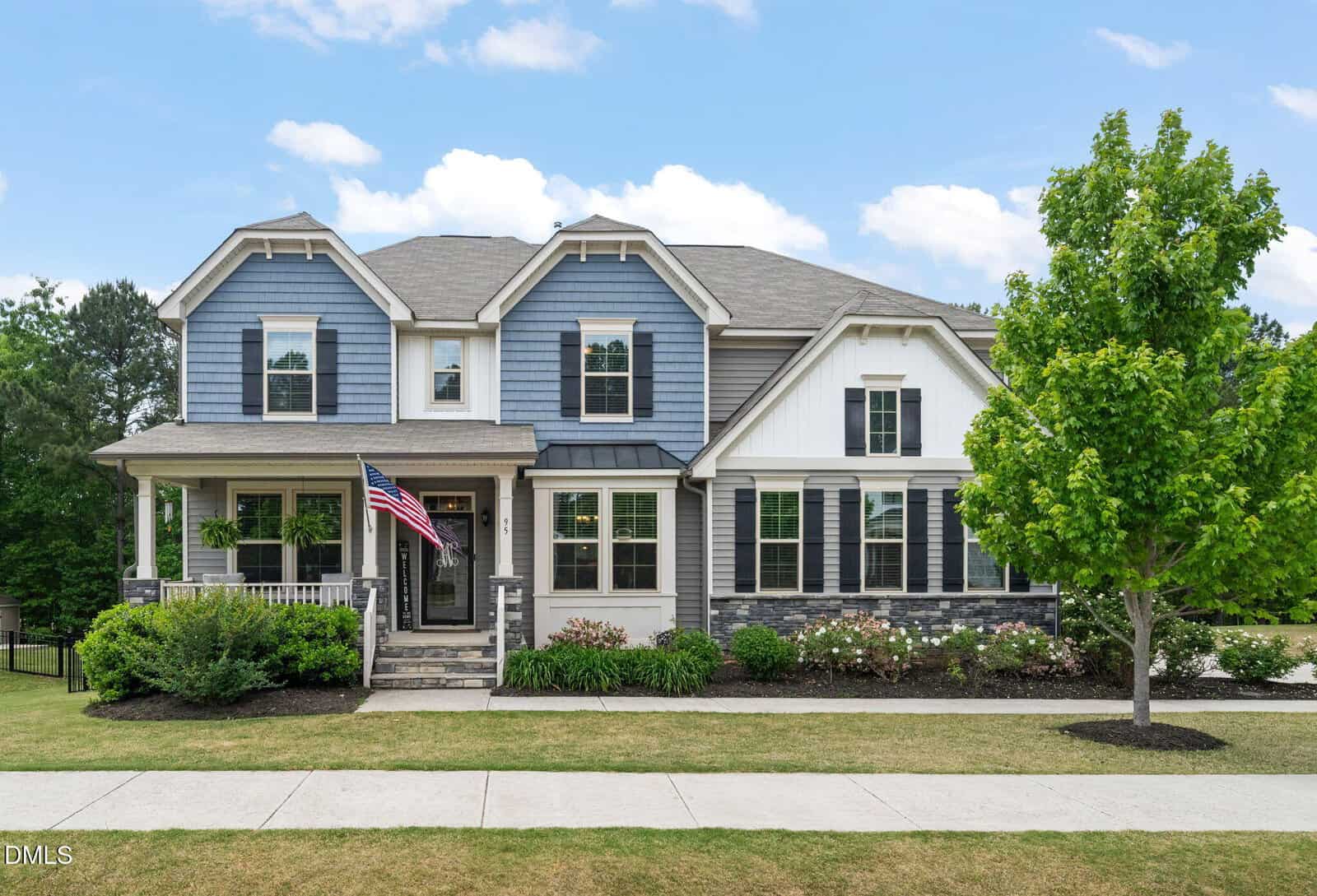Two-story suburban house with gray and white siding, black shutters, and a covered front porch. An American flag hangs by the entrance, with green bushes and a tree in the landscaped front yard.