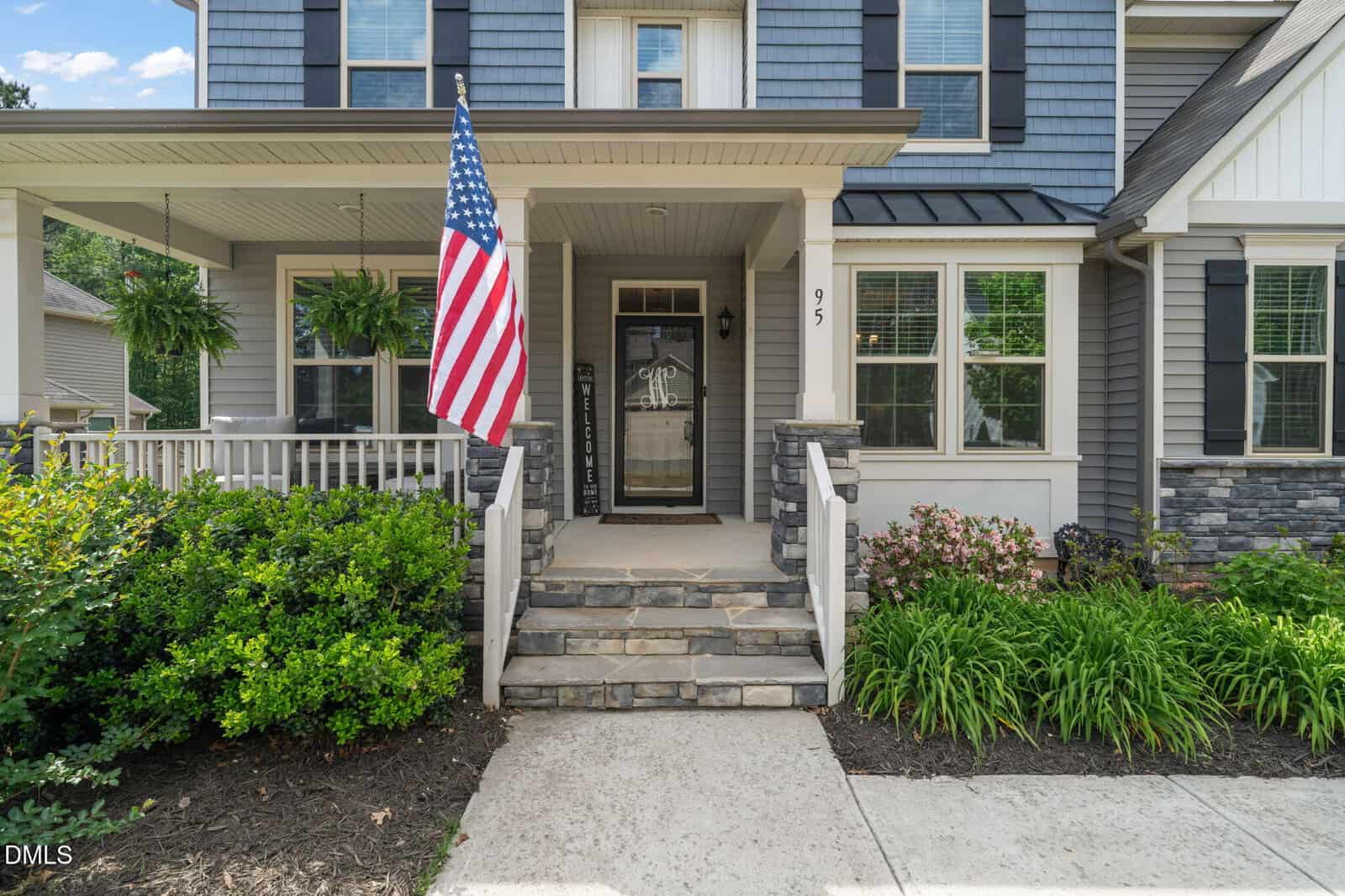 Front view of a gray house with white trim, stone accents, a porch with plants, steps leading to a glass door, an American flag hanging by the entrance, and landscaped greenery along the walkway.