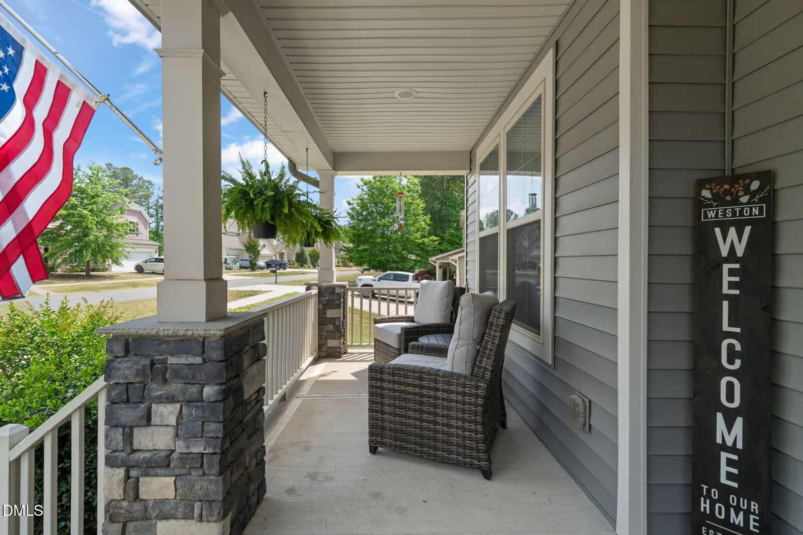 A cozy front porch with wicker chairs and cushions, gray siding, stone columns, a hanging fern, an American flag, and a "Welcome to our home" sign by the door on a sunny day.