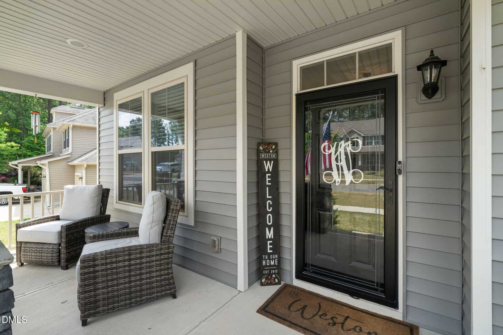 A cozy front porch with two cushioned chairs, a "Welcome" sign, a doormat, and a black storm door with decorative writing. The house exterior is gray siding with white trim and multiple windows.