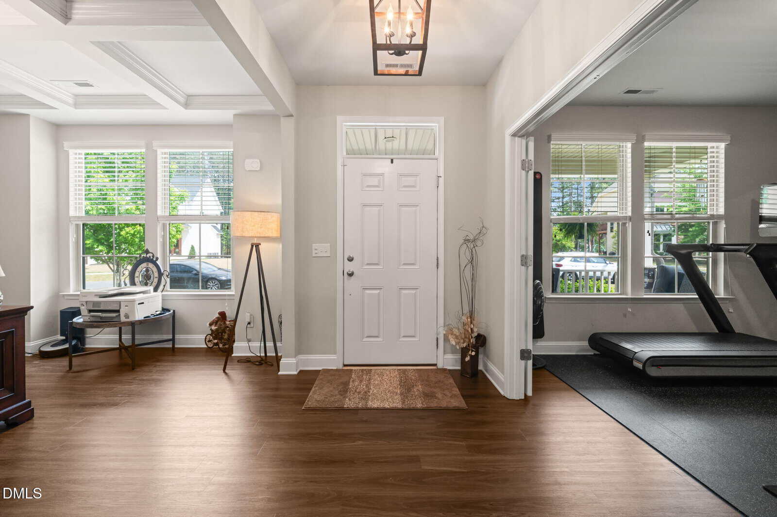 Bright entryway of a modern home with light walls, wood flooring, a central white front door, large windows, a treadmill in an adjacent room, and decorative items on a side table and in the corner.