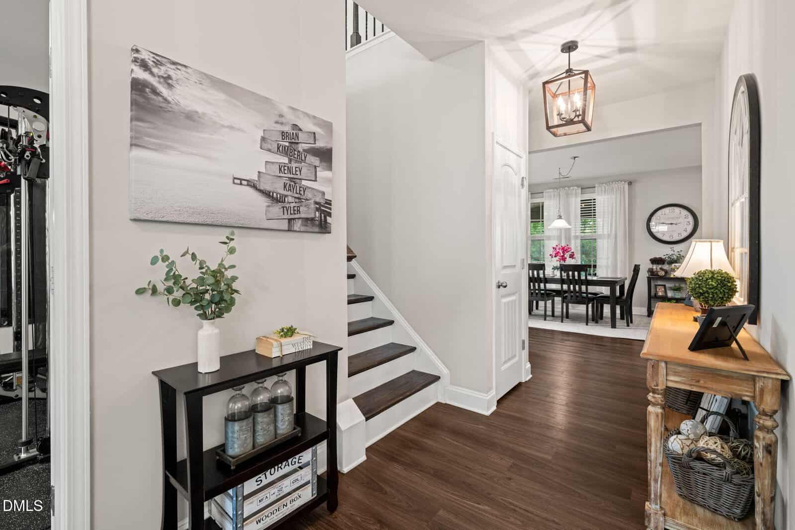 Bright hallway with dark wood floors, a staircase, small black shelf with plants, and a wooden console table. Dining area with flowers and a chandelier visible in the background. Black and white wall art hangs on the wall.
