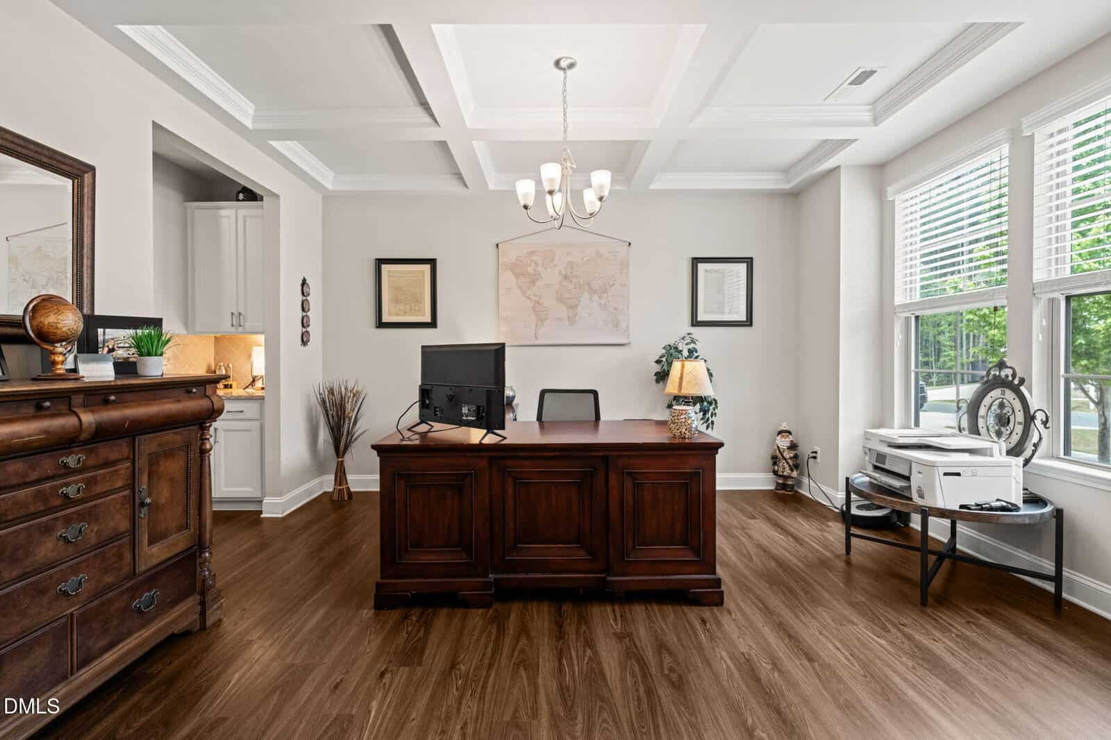 Elegant home office with a large wooden desk, computer, globe, and decor. A window lets in natural light. Wall art, chandelier lighting, and wood floors complete the room. A printer sits on a side table near the window.