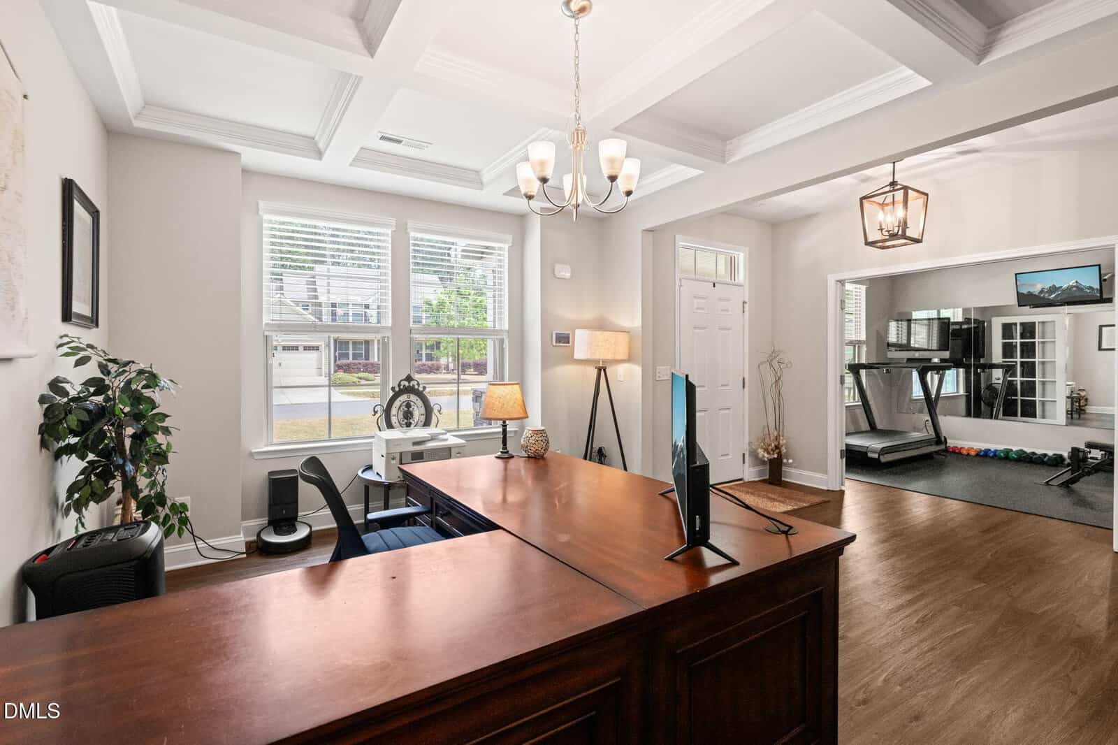 A bright home office with a wooden desk, computer monitor, lamp, and decor. Large windows let in natural light. An open doorway leads to a home gym with a treadmill and free weights.