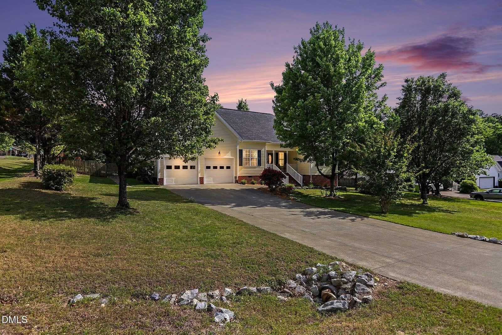 A single-story house with a front porch and two-car garage, surrounded by trees and a well-kept lawn, sits at the end of a long driveway at sunset with a colorful sky in the background.