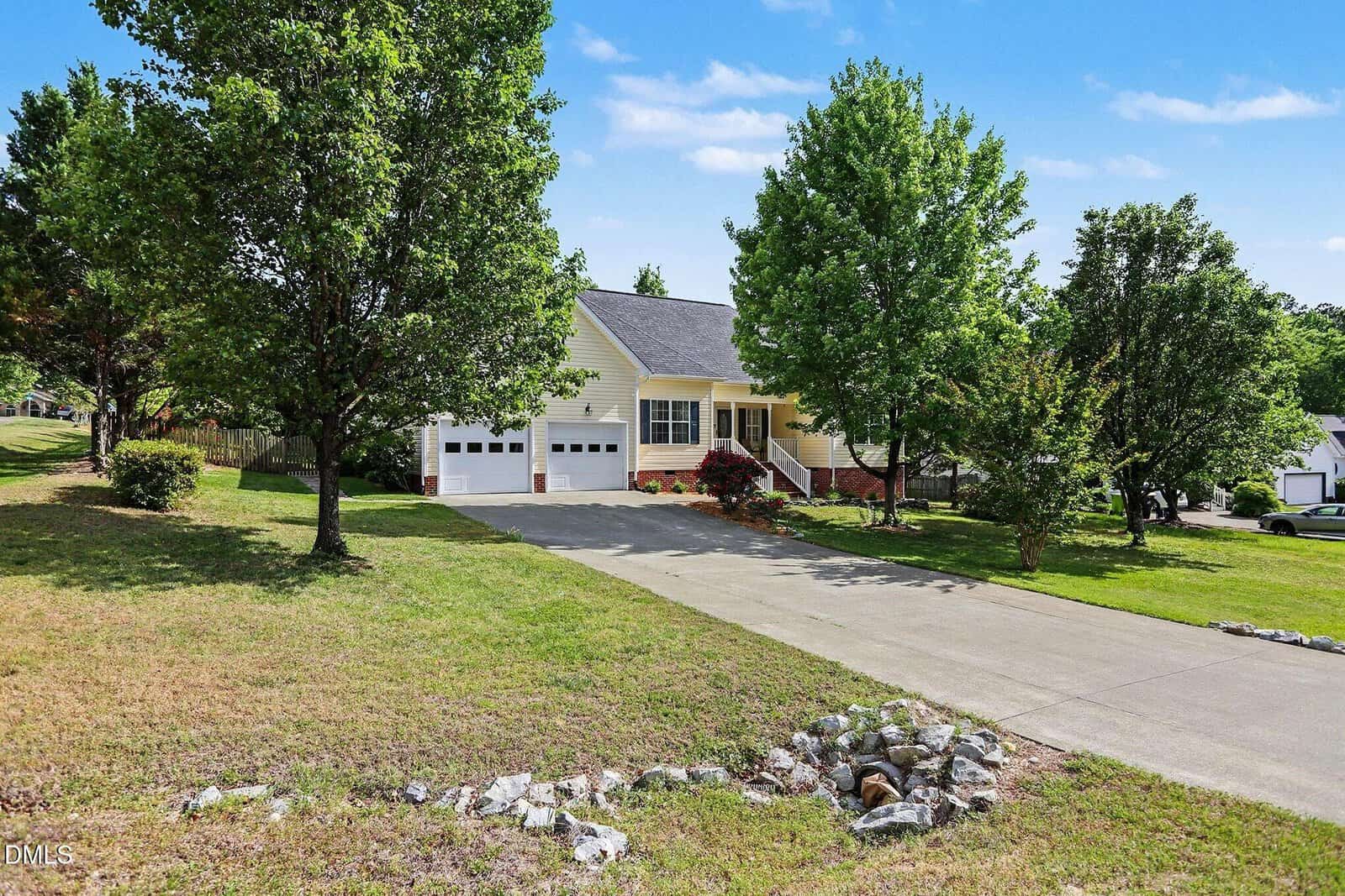 A yellow house with a front porch, two-car garage, and a long driveway, surrounded by green grass, mature trees, and landscaped rocks under a blue sky.