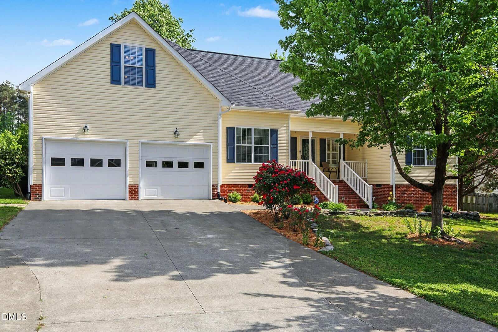 A beige house with two garage doors, white trim, and a front porch with steps. There are shrubs and a blooming red bush by the entrance, a large driveway, green grass, and trees in the yard under a blue sky.
