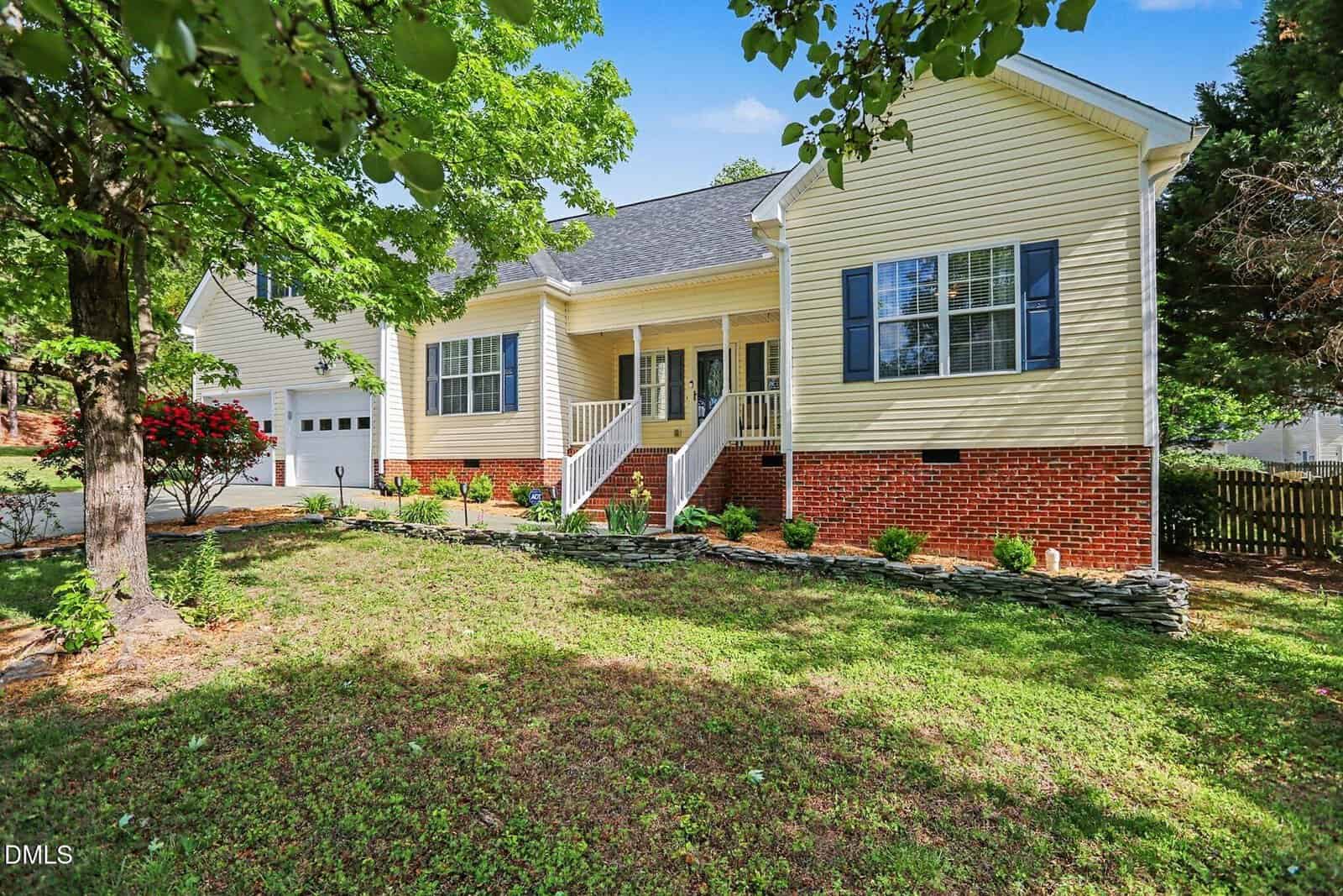 A yellow house with white trim and a brick foundation, featuring a front porch with white railings, a garage, and neatly landscaped shrubs set in a yard with large trees and green grass.
