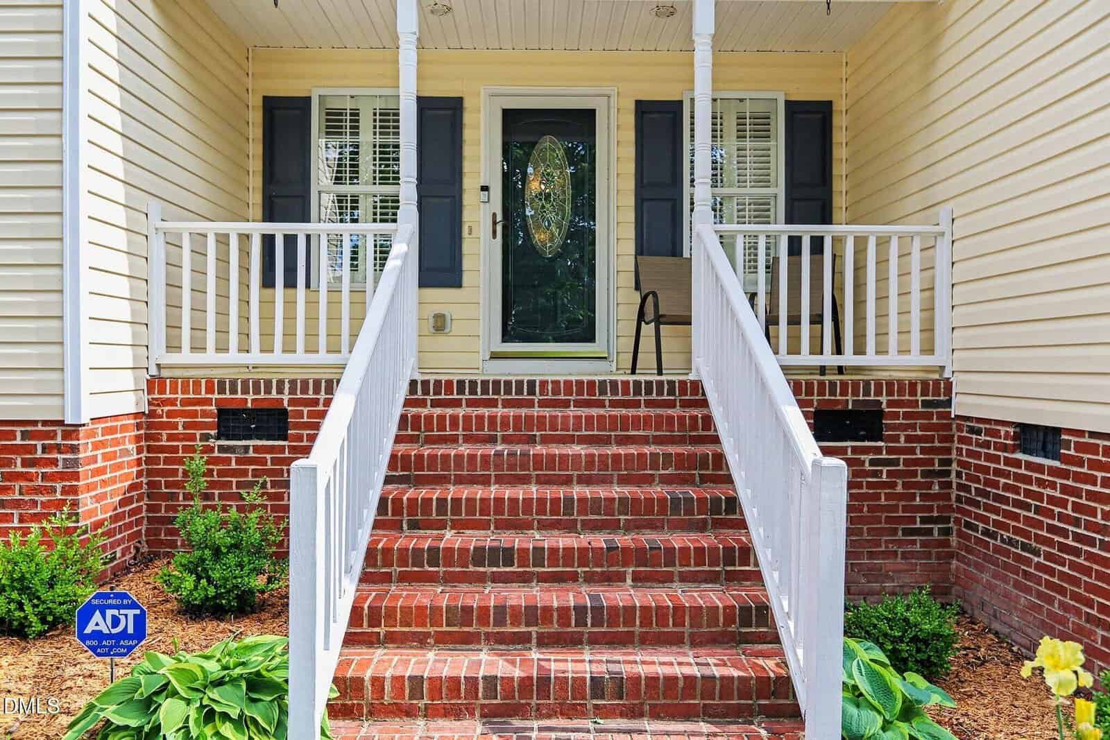 Front porch of a house with white railings, red brick steps, a black front door with glass panel, and two windows with shutters. Green plants are on either side of the steps. An ADT security sign is visible.
