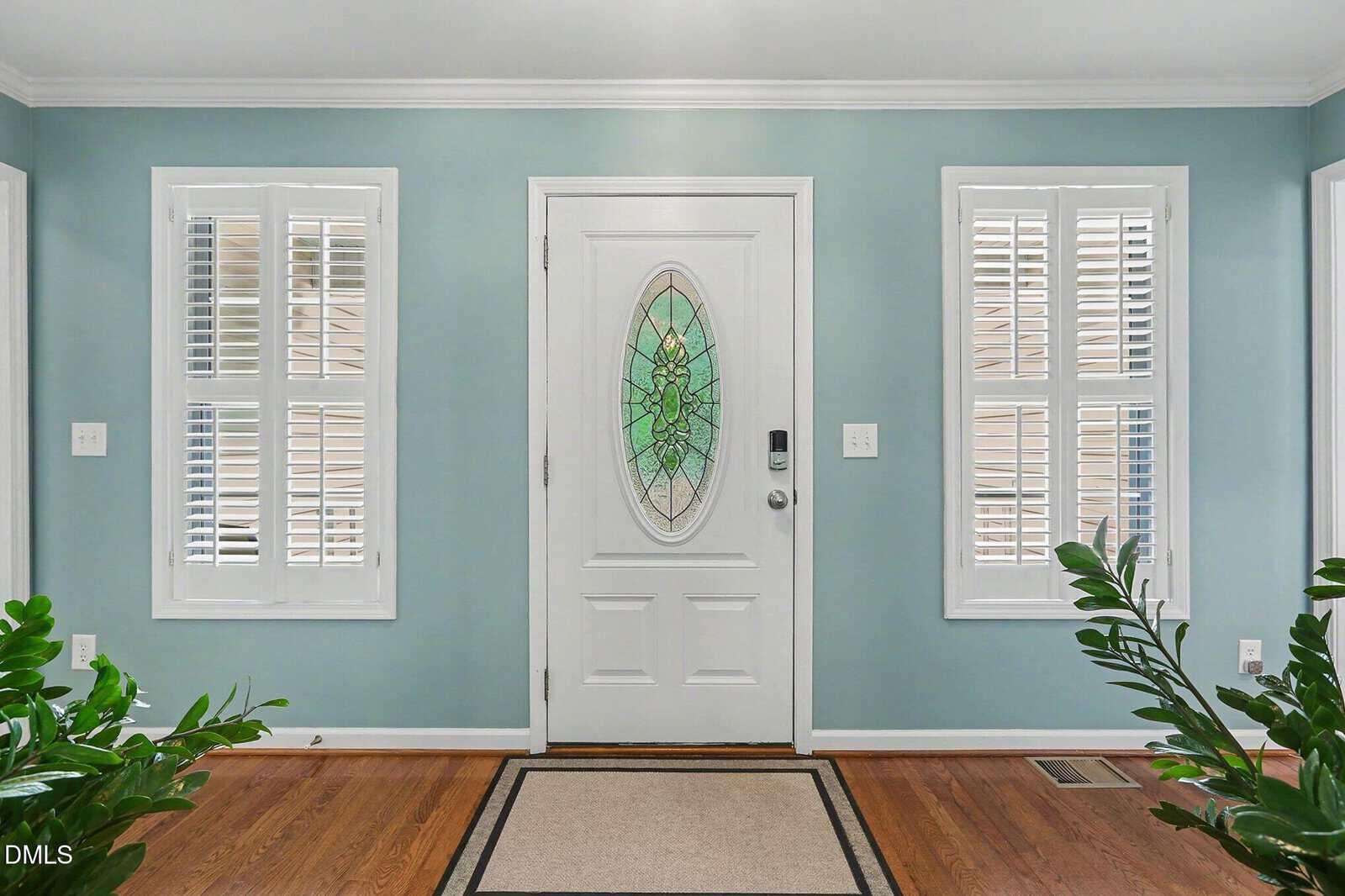 A white front door with an oval stained glass window is centered between two sets of double windows with white shutters. The walls are light blue, and green plants frame the entryway. A rug lies on the wooden floor.