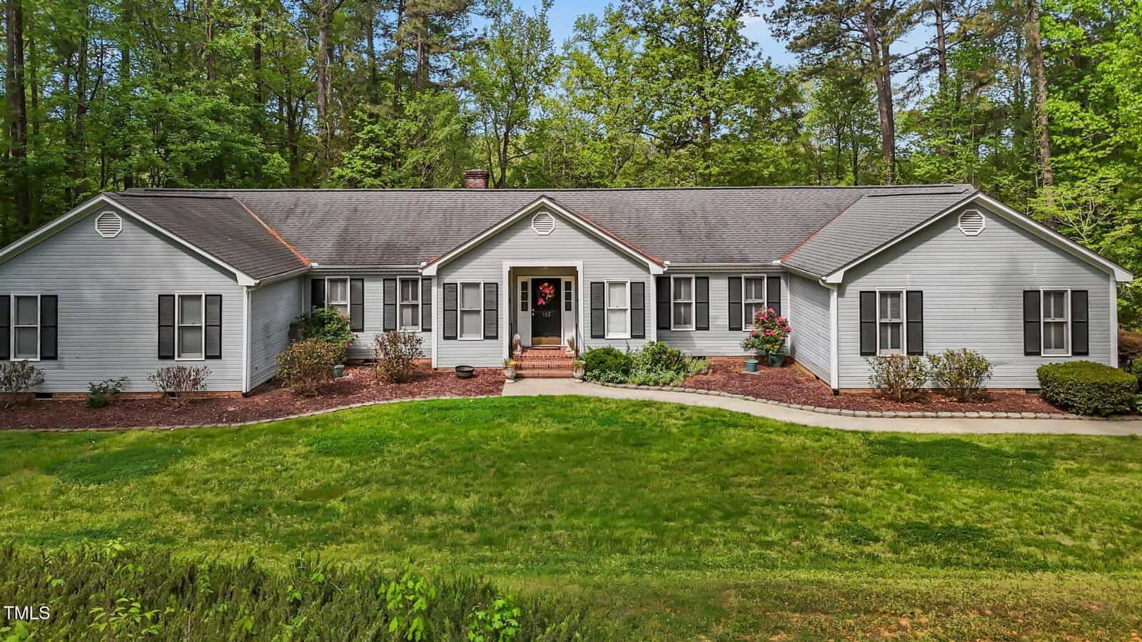 A single-story gray house with white trim and black shutters, surrounded by green grass, shrubs, and trees. There are steps leading to the front door, which is decorated with a wreath.