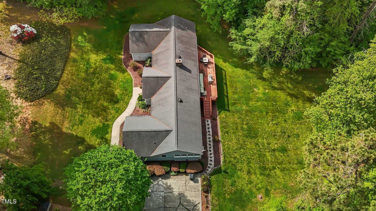 Aerial view of a single-story house with a gray roof, wooden deck, and driveway, surrounded by green grass, trees, and landscaped bushes in a spacious yard.