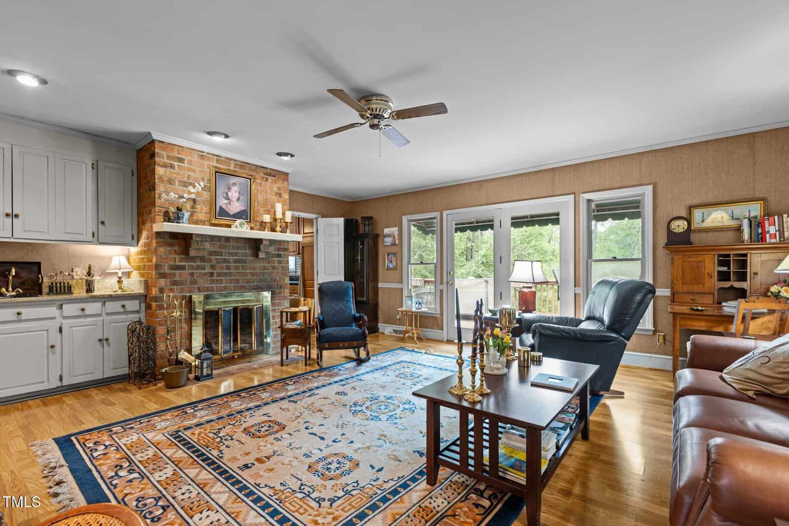 A cozy living room with a brick fireplace, armchairs, a large patterned rug, wooden floors, built-in shelves, and glass doors leading to a patio with greenery visible outside.