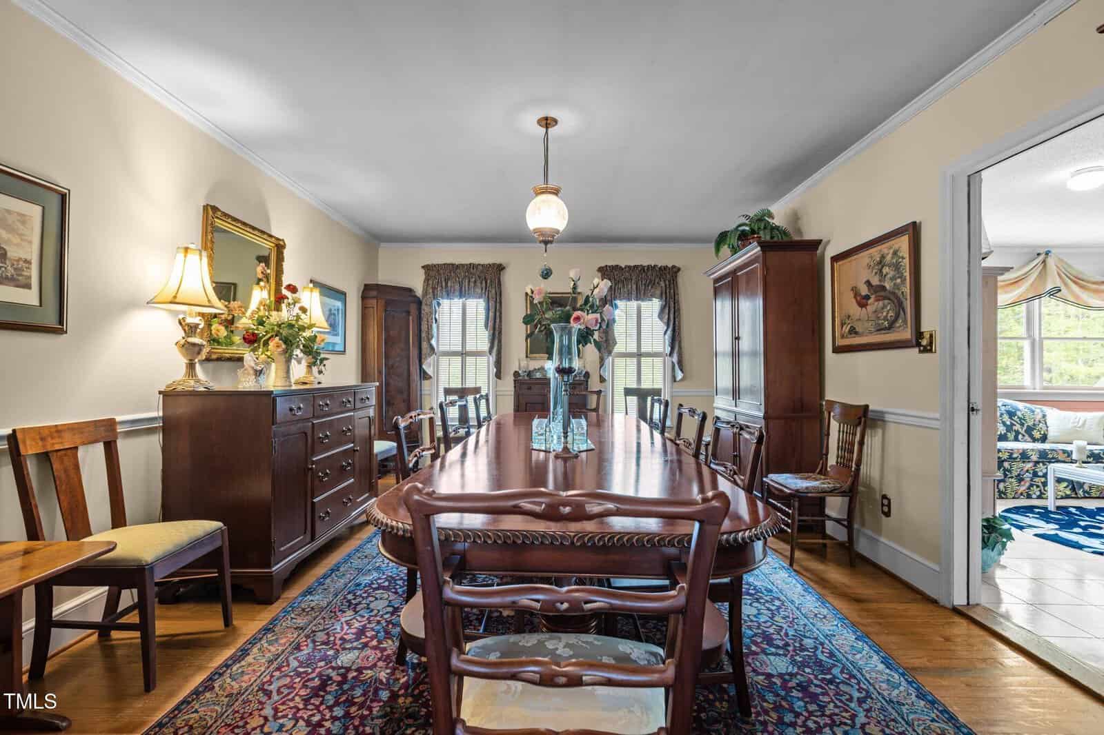A traditional dining room with a long wooden table, matching chairs, a sideboard with lamps and flowers, a china cabinet, framed art, and windows with curtains. A doorway leads to another room with a sofa.