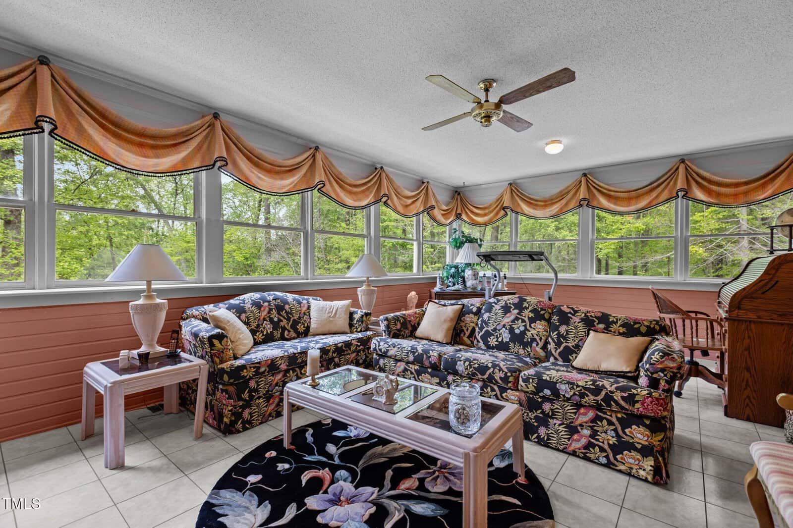A bright sunroom with large windows, floral-patterned sofas, glass coffee tables, lamps, a ceiling fan, and exercise equipment. Green trees are visible outside, and valance curtains decorate the windows.