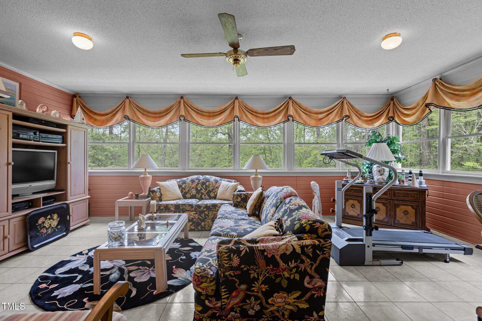 Bright sunroom with floral sofas, a glass coffee table, TV, treadmill, lamps, and large windows with orange valances. Green trees are visible outside, and light fills the tiled room.