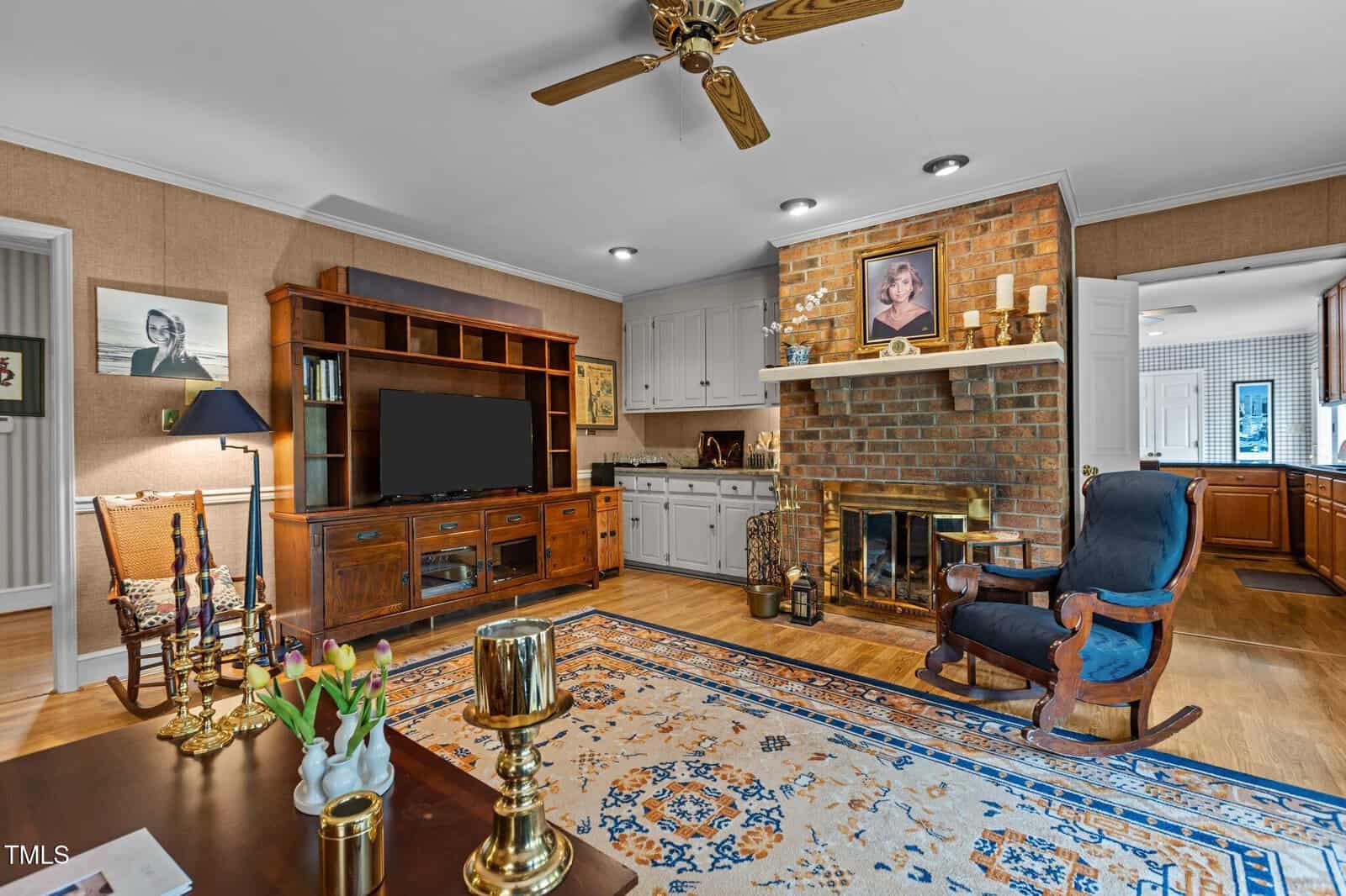 A cozy living room with a brick fireplace, TV stand, rocking chair, ceiling fan, and a patterned rug. Photos and decor items are on the walls and shelves, and a dining area is visible to the left.