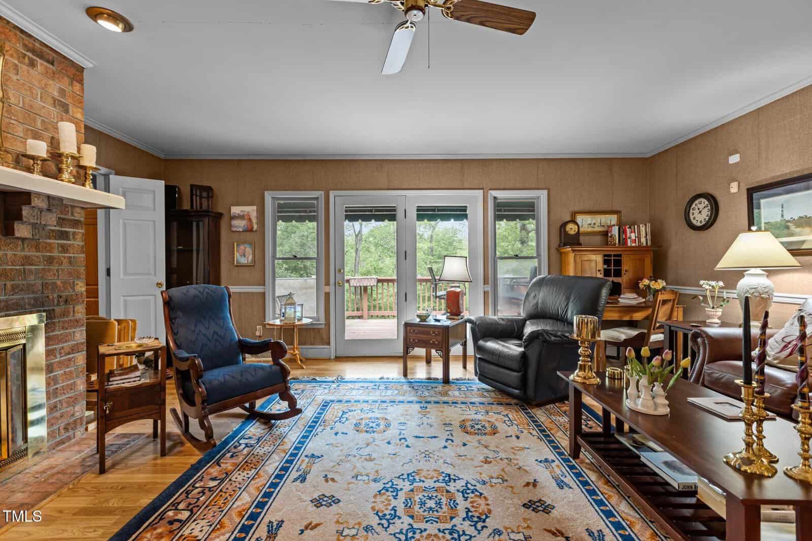 A cozy living room with wooden floors, patterned rug, brick fireplace, blue armchair, black recliner, bookshelves, and glass doors opening to a deck with outdoor views. Lamps and decor add warmth to the space.