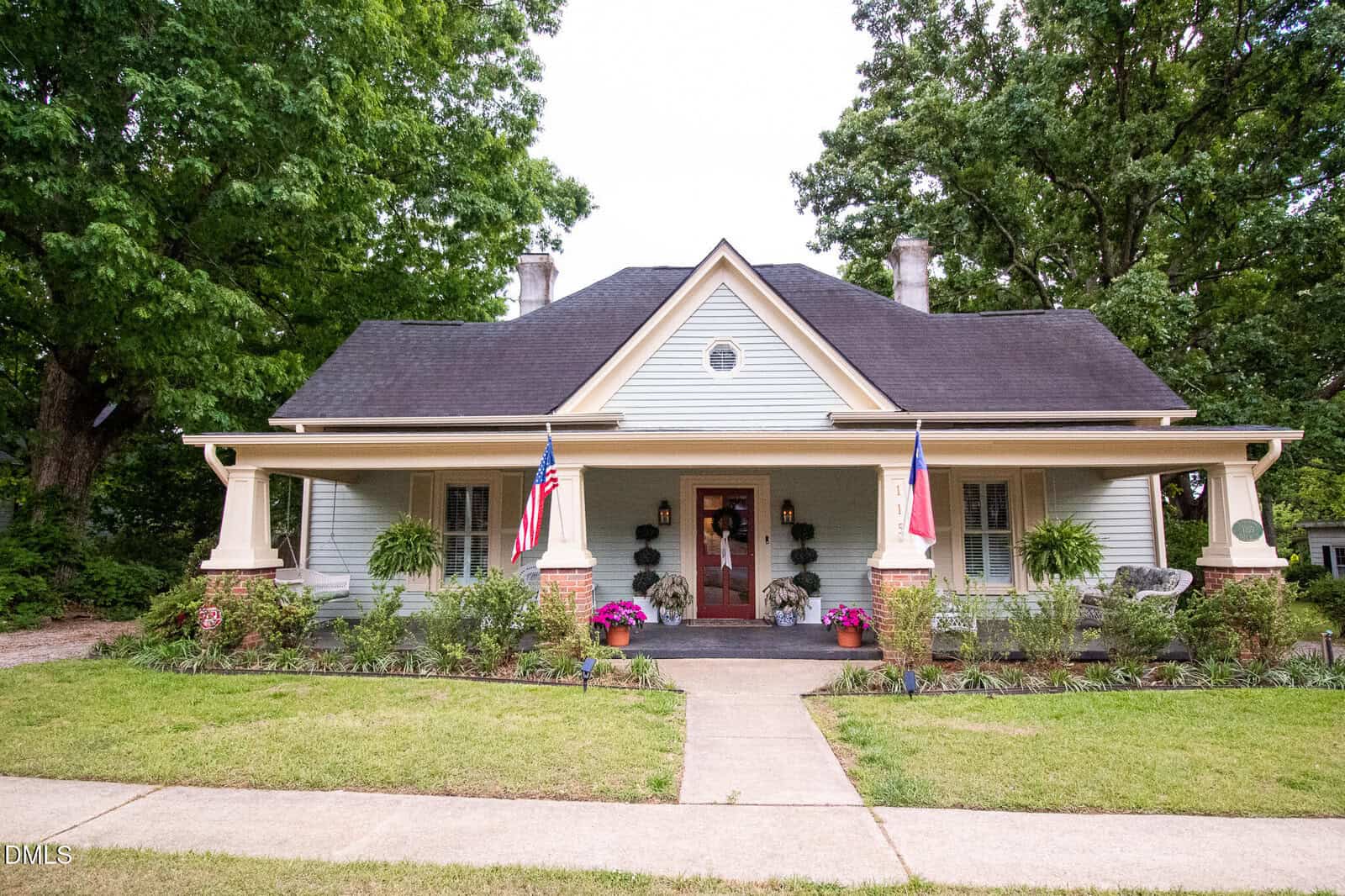 A charming light green house with a wide front porch, two American flags, potted plants, and a tidy lawn. Brick columns frame the porch, and large trees surround the home.