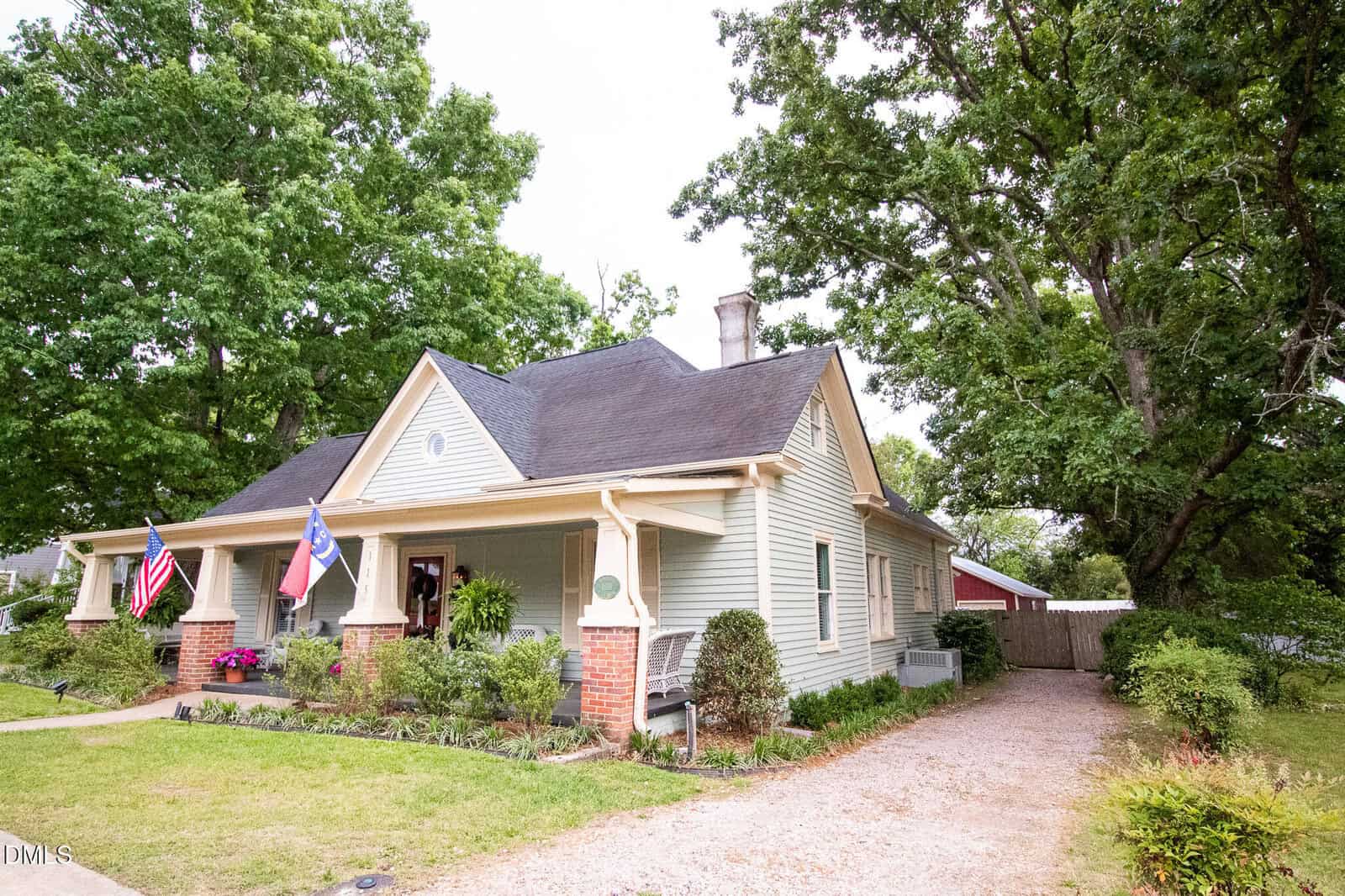 A light green, single-story house with a covered front porch, two flags displayed, and a gravel driveway. The yard is landscaped with shrubs and trees surround the home.