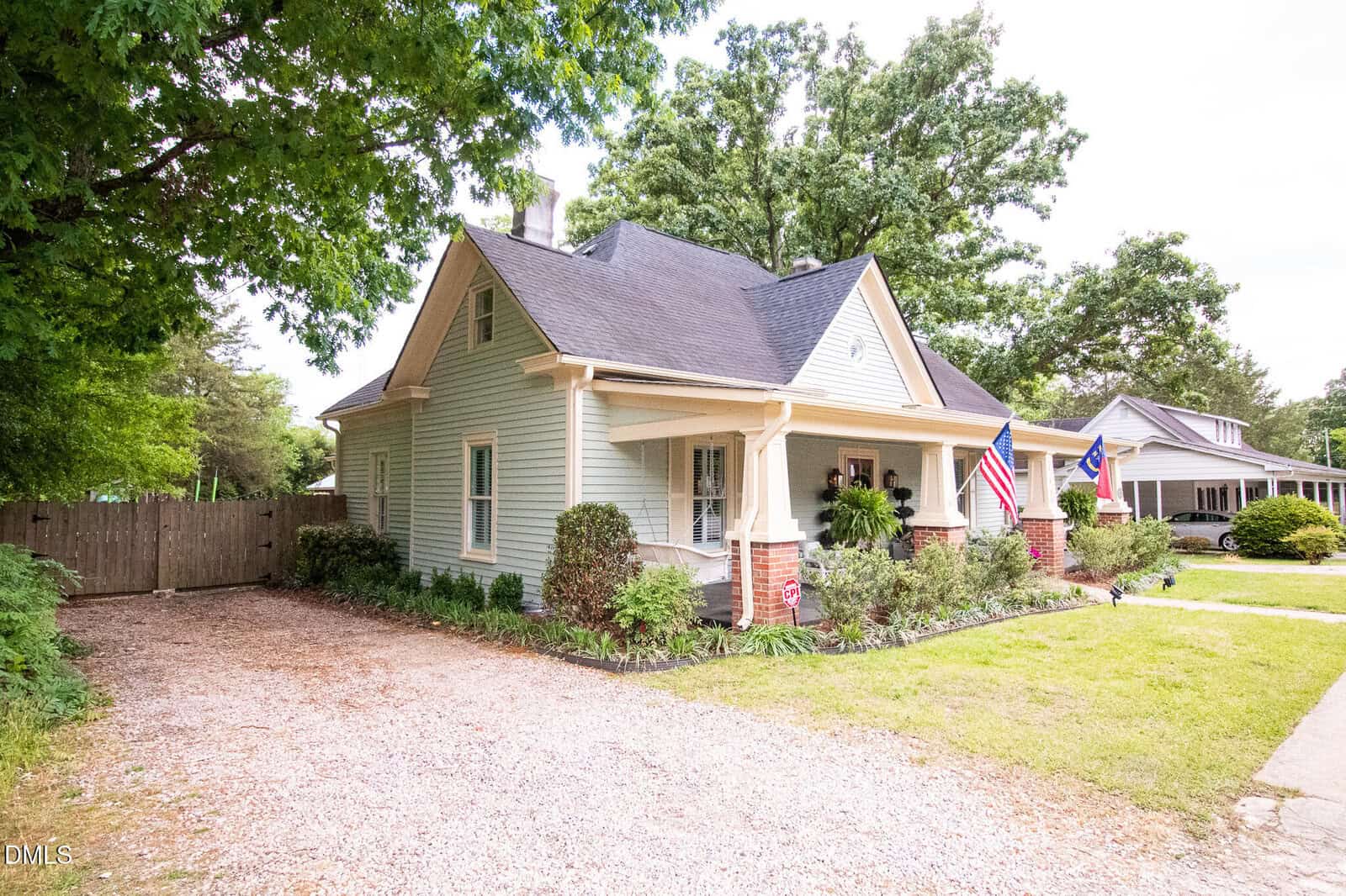 A light green house with a steep gabled roof, a covered front porch, American and state flags, brick pillars, garden beds, and a gravel driveway, surrounded by trees and neighboring houses on a sunny day.