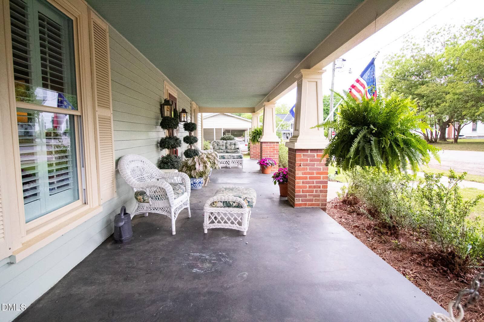 A spacious front porch with pale green walls, white wicker chairs, potted plants, hanging ferns, and an American flag, overlooking a yard and quiet street.