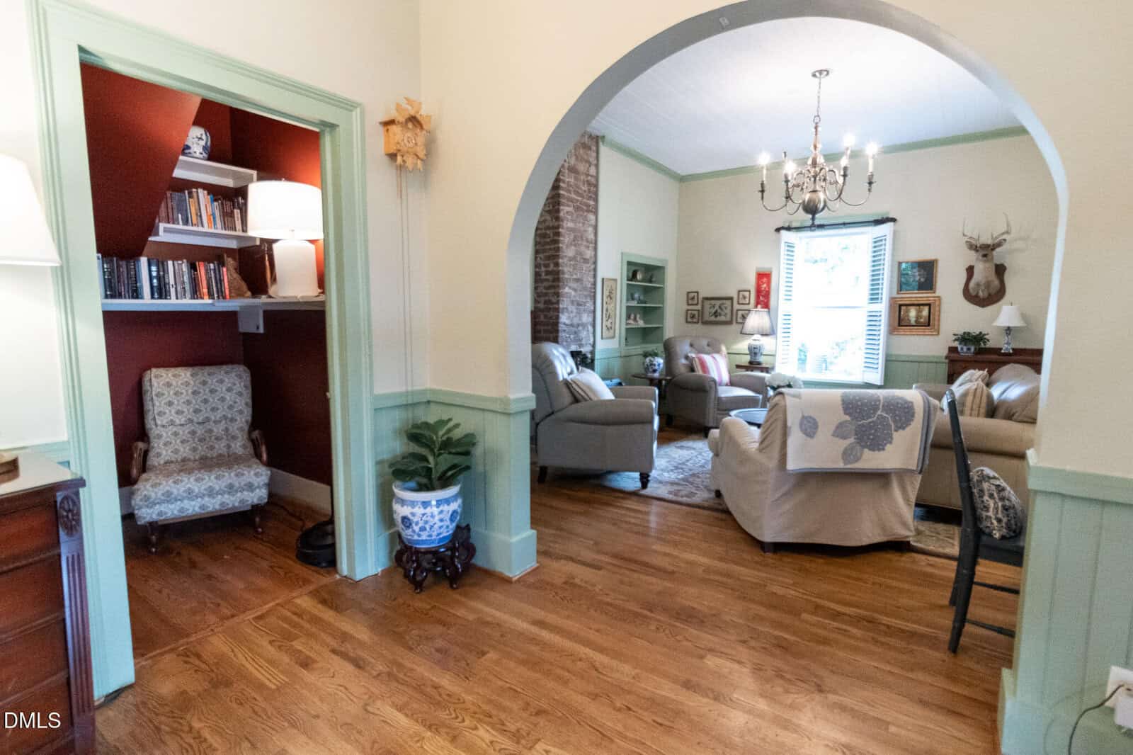 A cozy living room with hardwood floors, neutral sofas, and a chandelier. An archway separates a reading nook with a patterned chair, bookshelves, and a lamp. There is also a potted plant and wall decor throughout the space.
