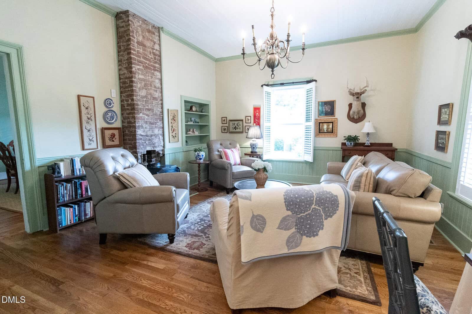 Cozy living room with beige armchairs, sofa, and a floral quilt. Exposed brick runs up one wall. Shelves, artwork, and a deer head mount add charm. A window with shutters lets in natural light.