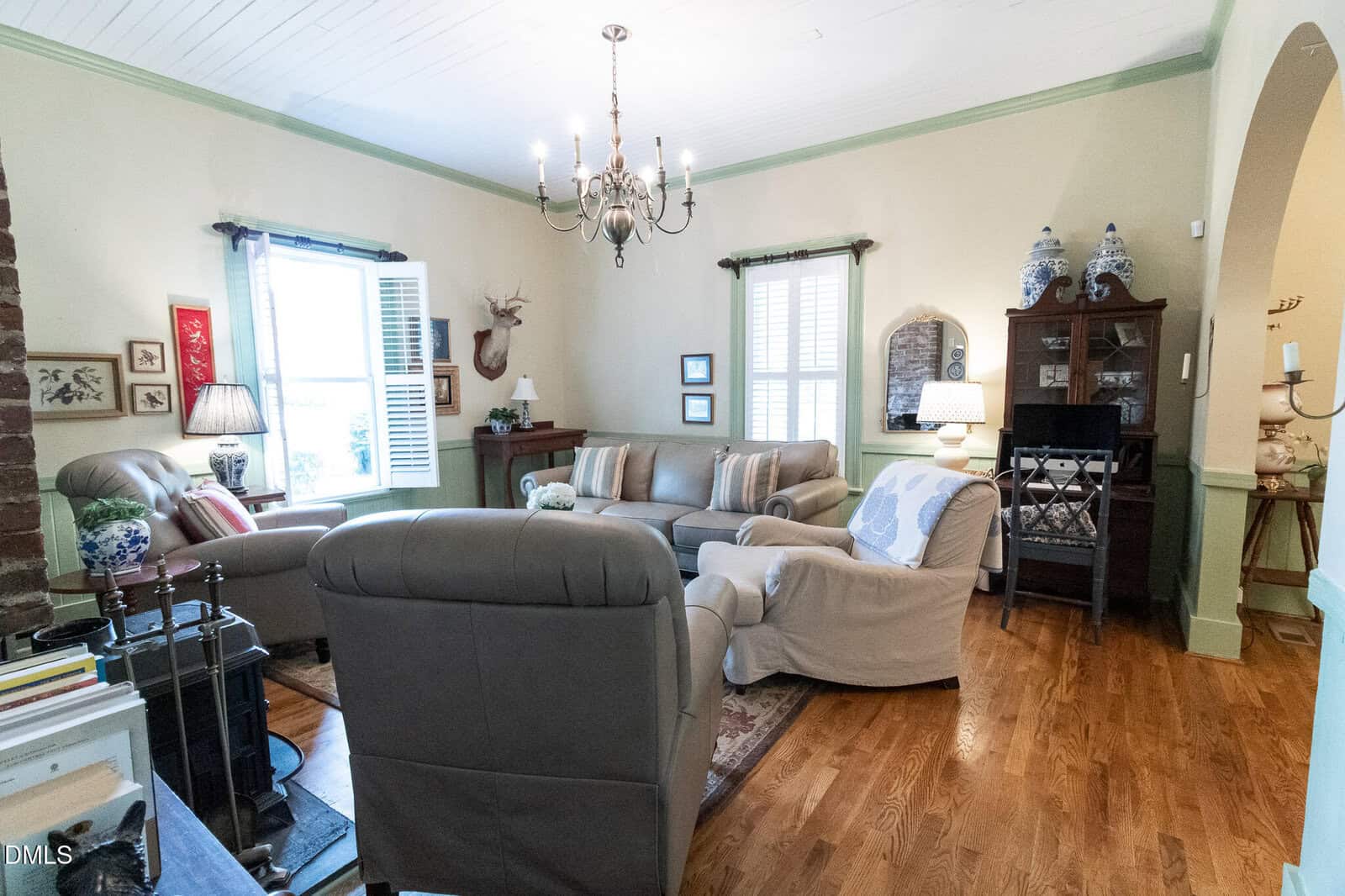 Cozy living room with neutral-colored sofas and chairs, hardwood floors, a chandelier, decorative wall items, a small cabinet, and windows with white shutters letting in natural light.