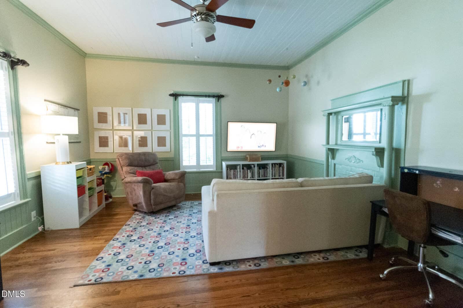 Bright living room with a sofa, recliner, TV, colorful rug, kids’ storage bins, framed art, fireplace, ceiling fan, and a small desk with a chair. Hardwood floors and mint green trim accent the space.