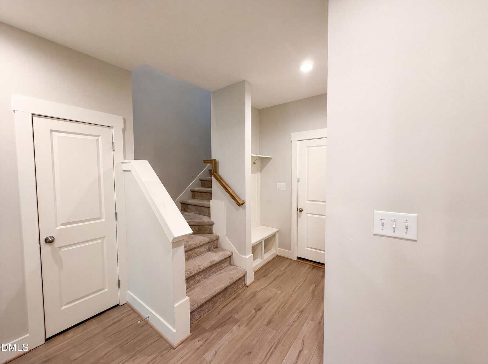 Modern entryway with light wood floors, white doors, carpeted stairs with a wood handrail, and a small built-in bench with hooks beneath a shelf. Walls are painted light gray, and a switch plate is visible.