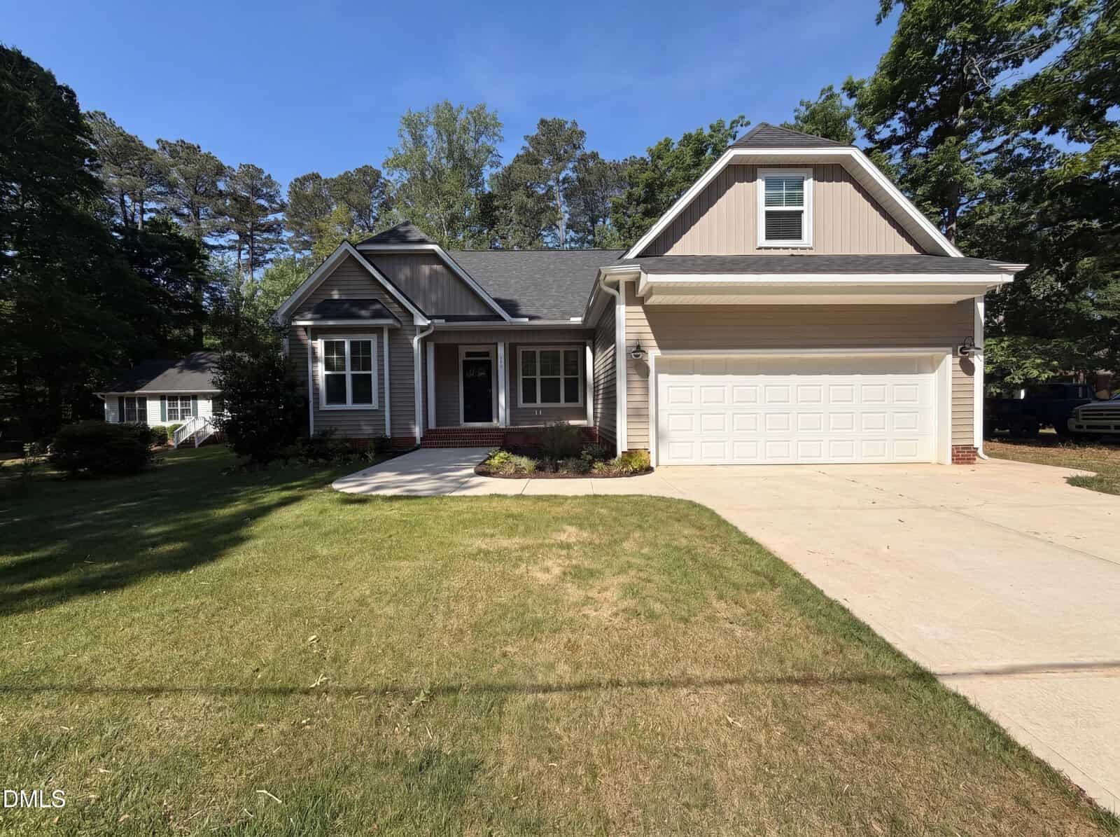 A modern single-story house with beige siding, a gray roof, white trim, large windows, and a two-car garage, set on a grassy lawn with trees and a driveway under a clear blue sky.
