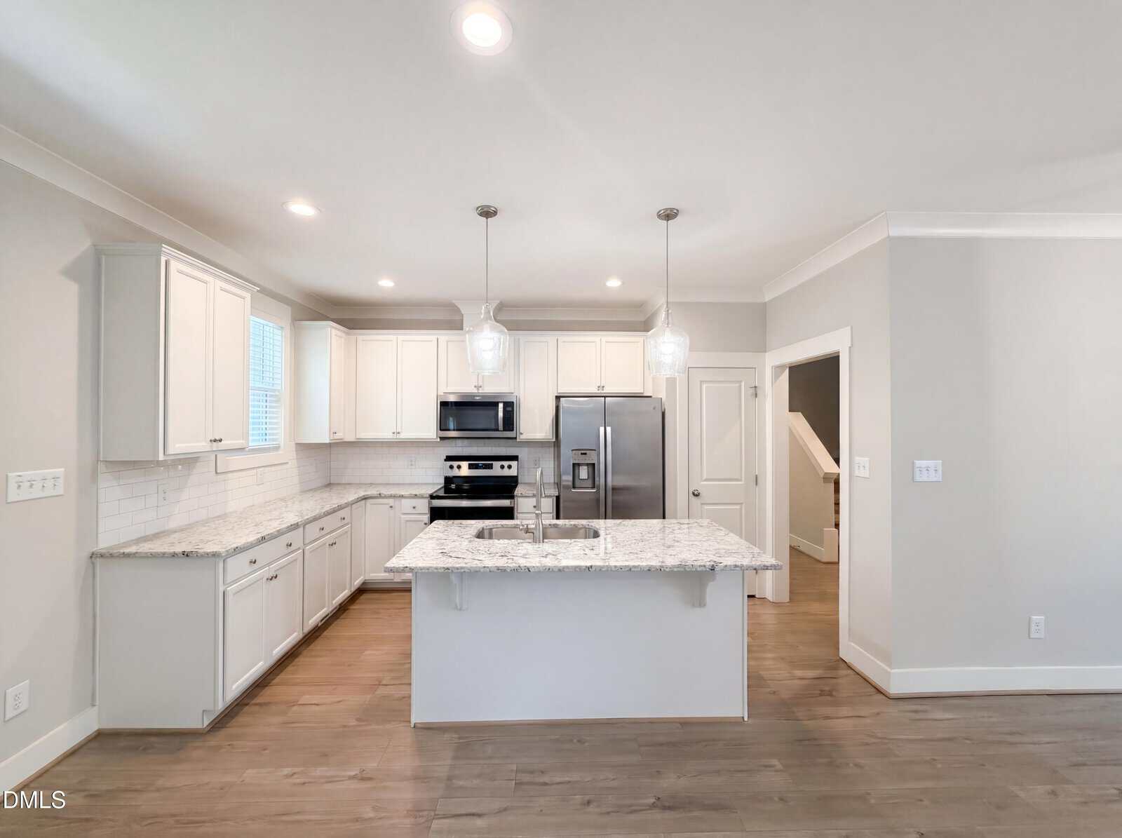 Modern kitchen with white cabinets, stainless steel appliances, a center island with sink, granite countertops, pendant lights, and wood flooring. A staircase and door are visible in the background.