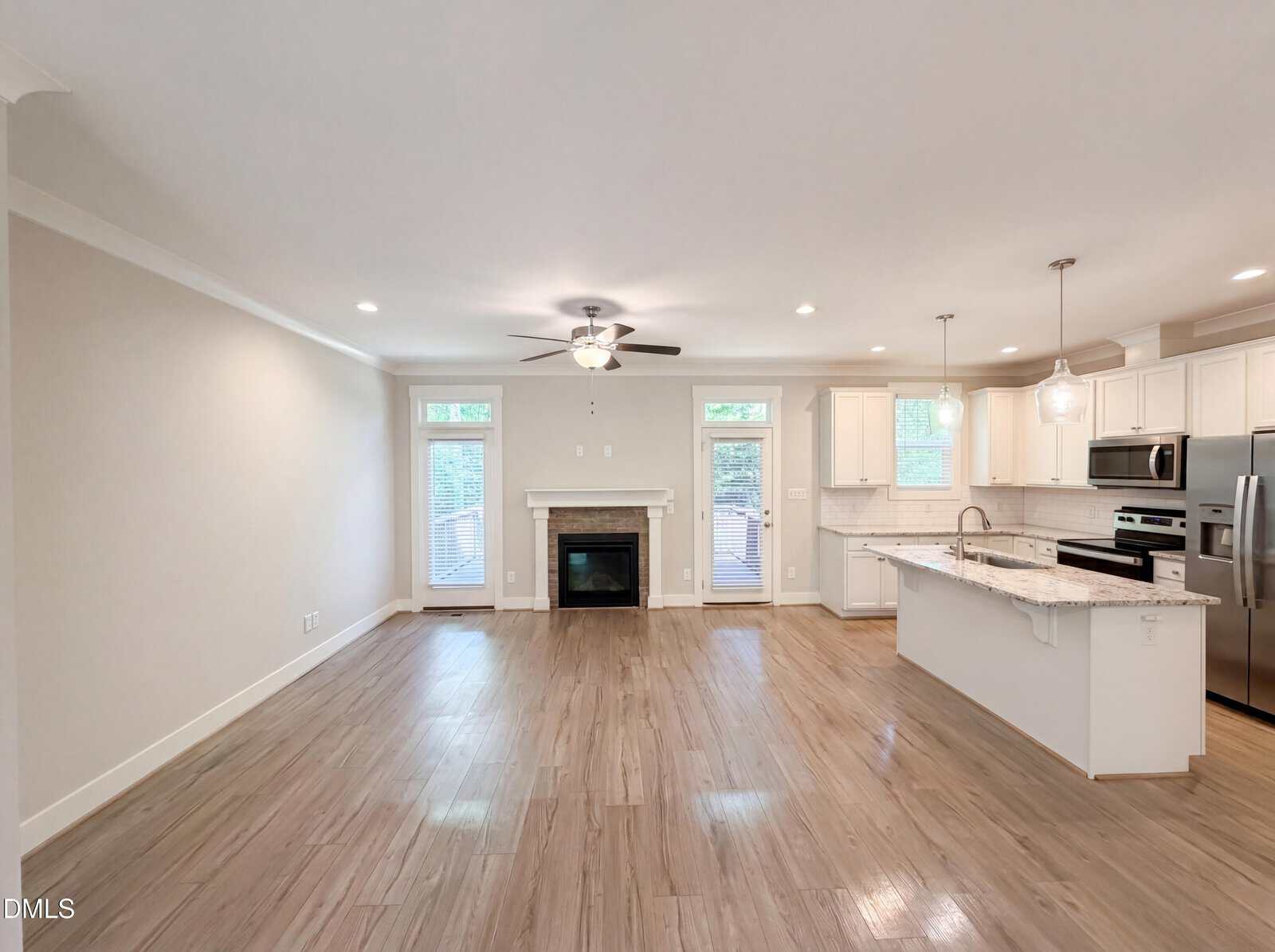 Open-concept living area with light wood floors, a ceiling fan, fireplace centered between two tall windows, modern white kitchen with island, stainless steel appliances, and pendant lighting.