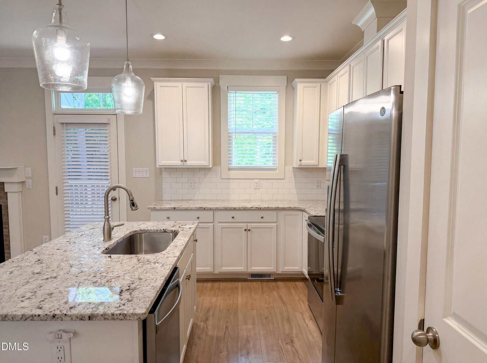 Modern kitchen with white cabinets, stainless steel appliances, marble countertops, a double sink on a center island, pendant lights, wood flooring, a window above the sink, and a door with blinds leading outside.