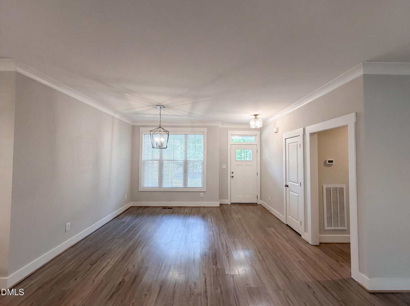 A spacious, empty living room features light gray walls, wood flooring, two ceiling lights, a large front window with blinds, and a white front door with side panels.