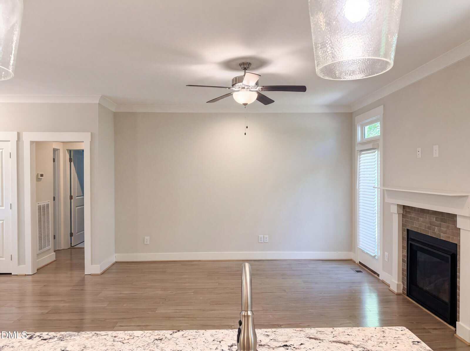 A bright, empty living room with wood floors, light gray walls, a ceiling fan, a fireplace with a white mantel, and large windows allowing natural light in. A kitchen counter with a sink is visible in the foreground.