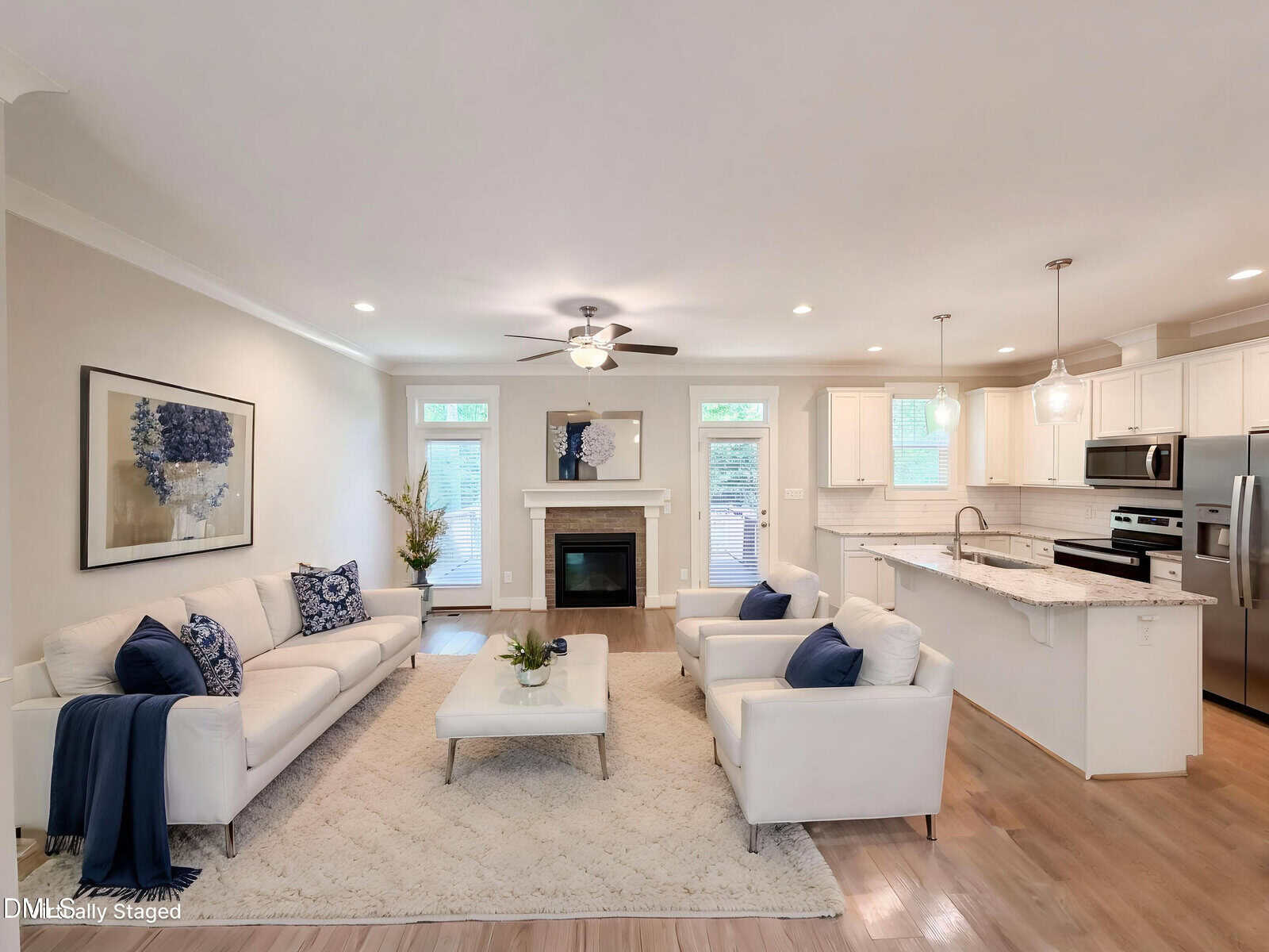A bright, modern living room with white sofas, blue accent pillows, a coffee table, a fireplace, large windows, and an open kitchen with white cabinets and stainless steel appliances.