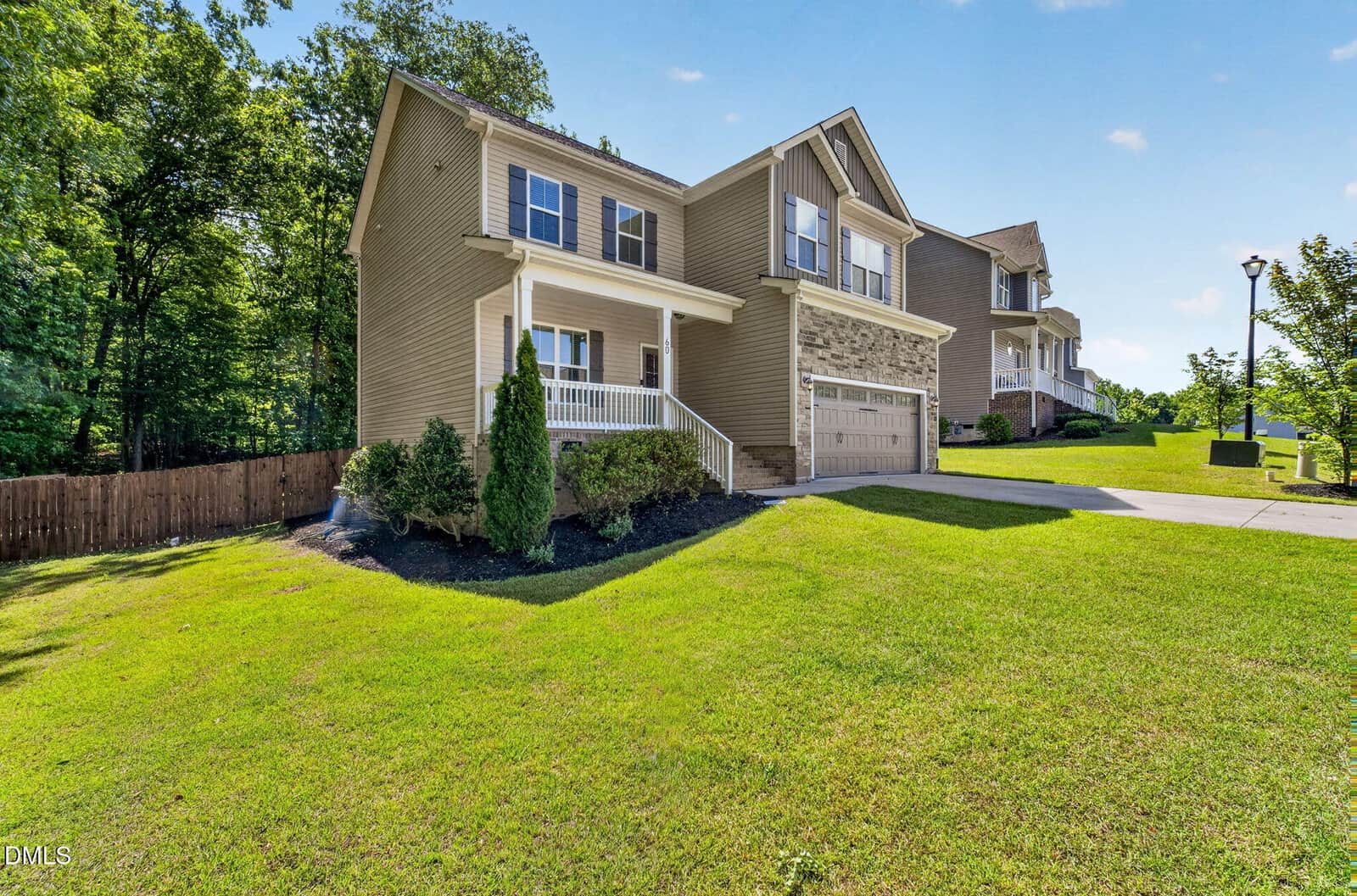 Two-story beige house with a front porch, double garage, and stone accents, set on a sloped green lawn with shrubs and a wooden fence, surrounded by trees and neighboring homes on a sunny day.