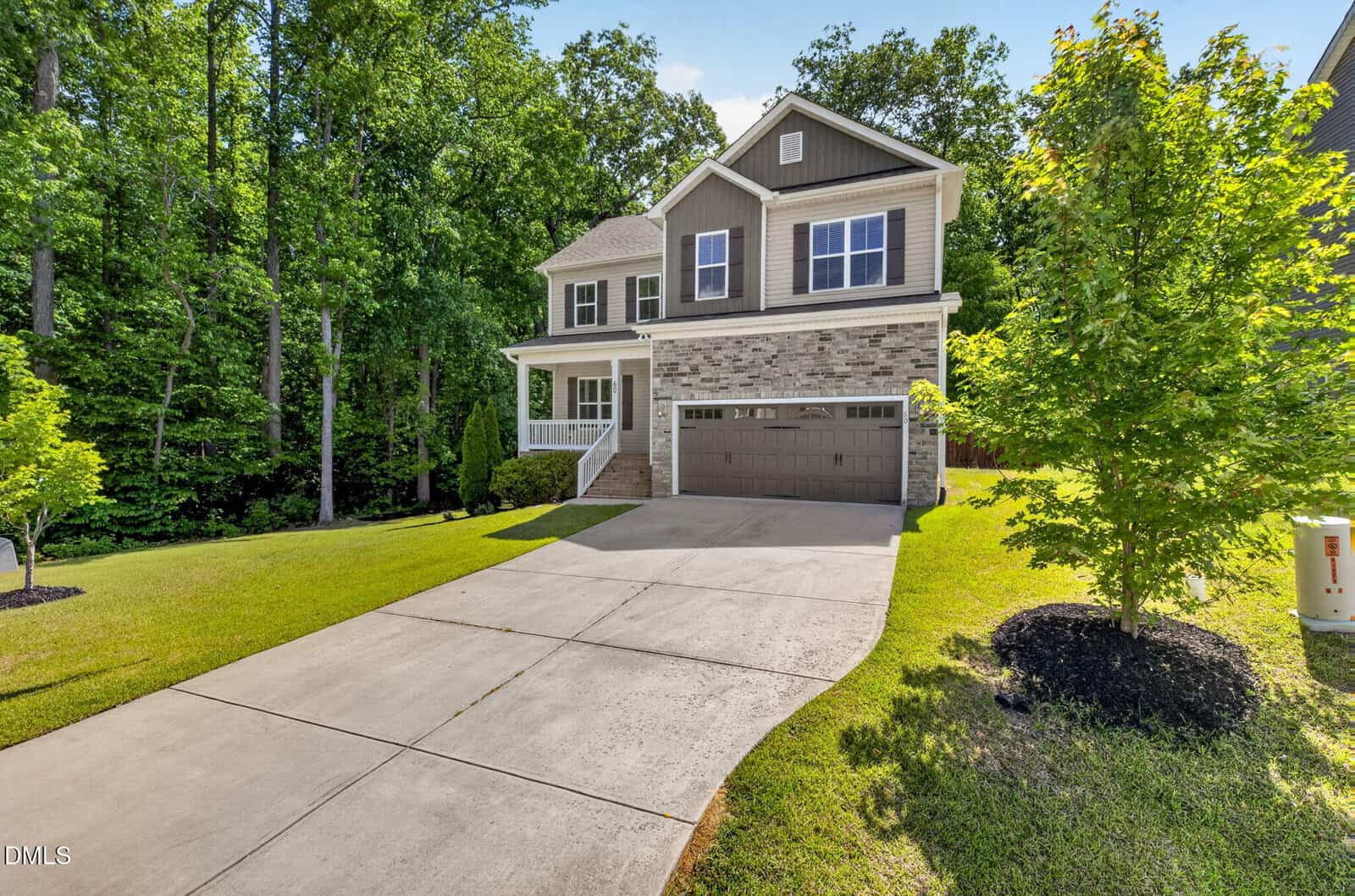 A modern two-story house with gray siding, stone accents, a covered front porch, and a double garage, surrounded by green grass and trees on a sunny day.