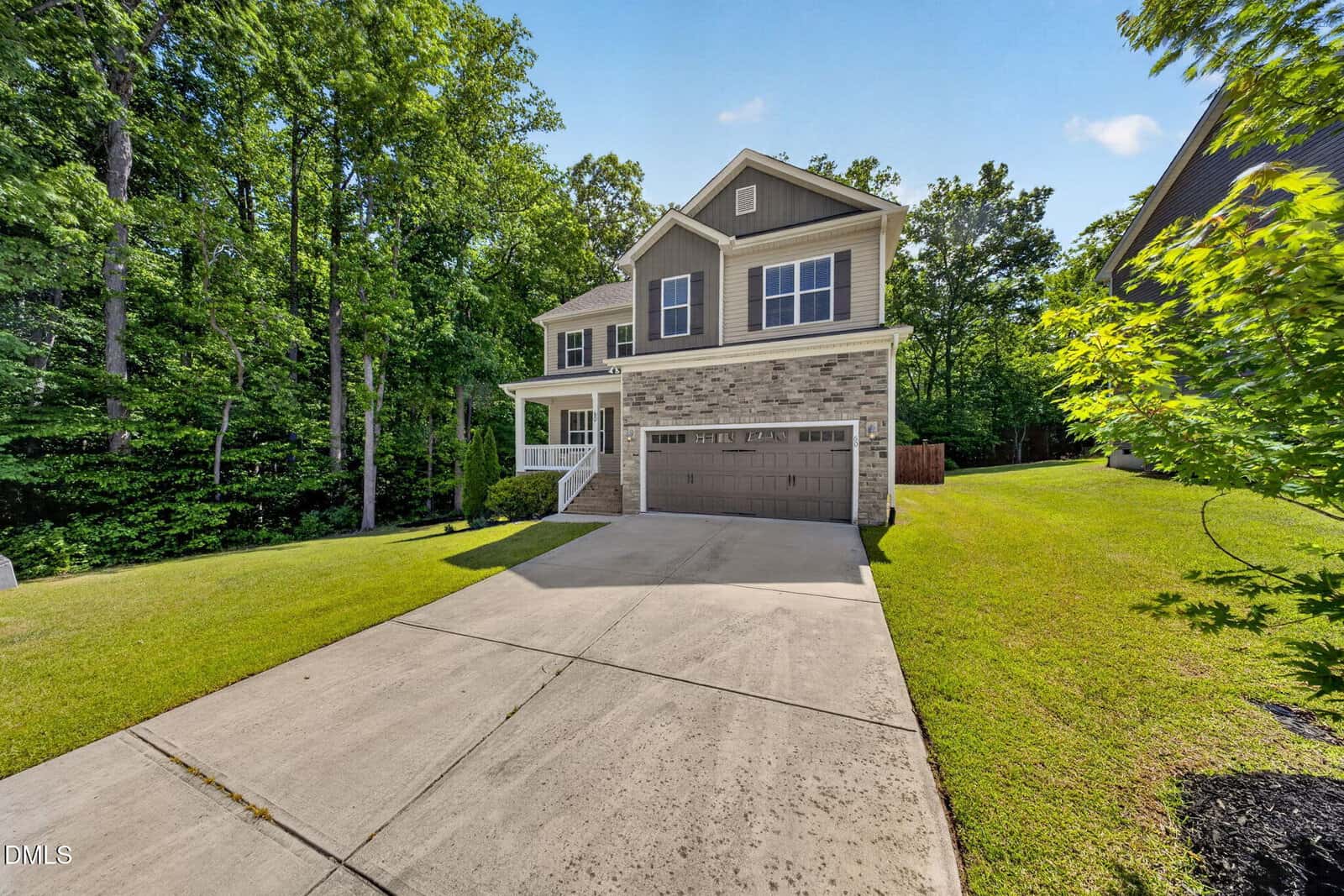 Two-story suburban house with gray and brick exterior, double garage, and white trim, set on a green lawn with a concrete driveway, surrounded by trees and blue sky.