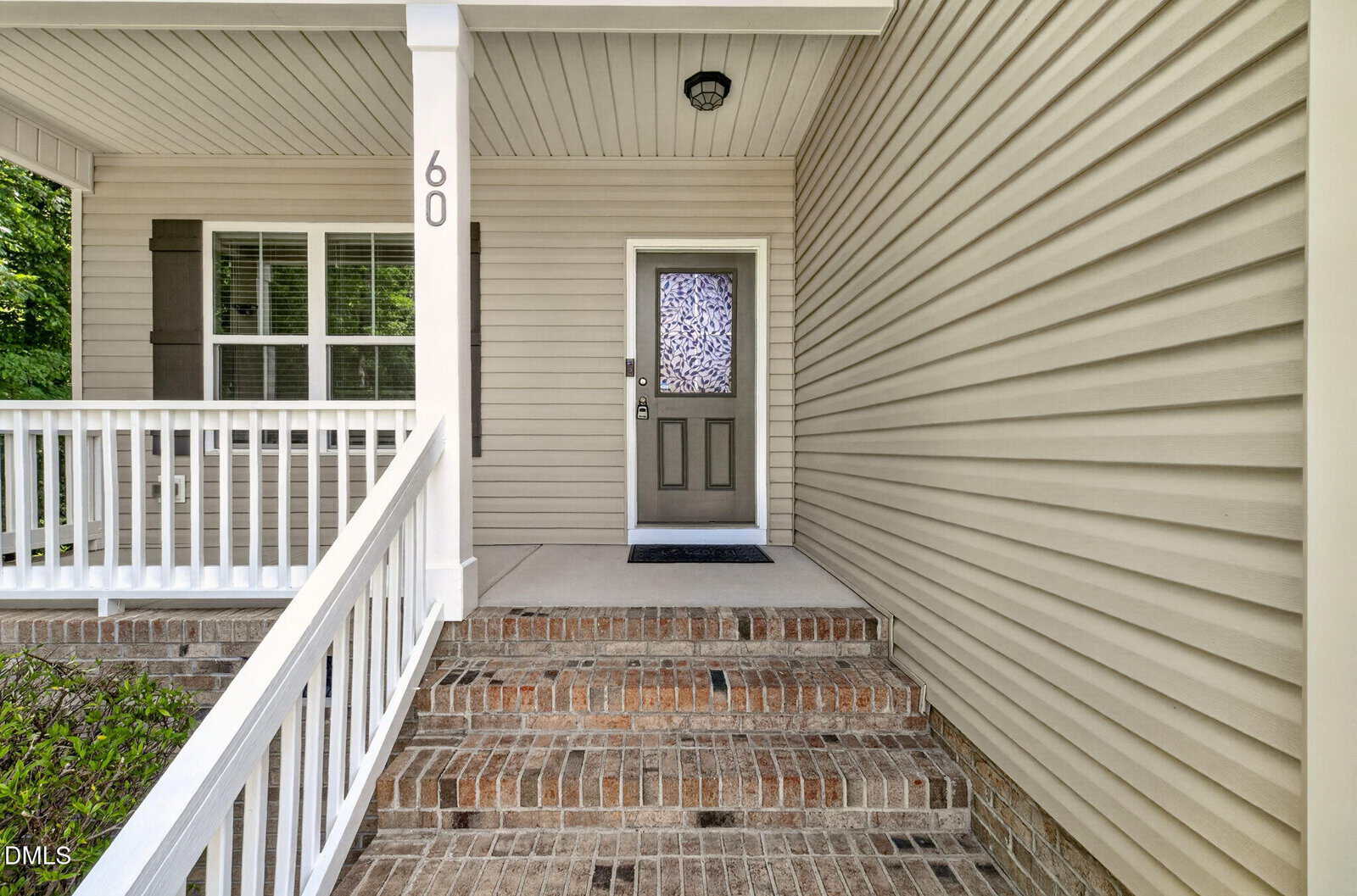 A beige house with horizontal siding, a covered front porch, white railing, brick steps, a dark green front door with a window, and the number 60 on a white post.