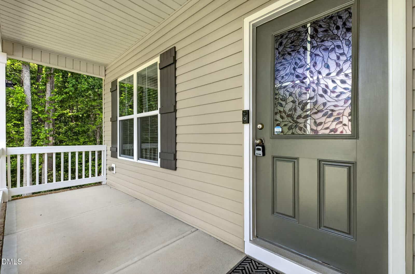A close-up view of a house’s front porch, featuring beige siding, a decorative glass front door, white railing, and double windows with dark shutters. Trees are visible in the background.