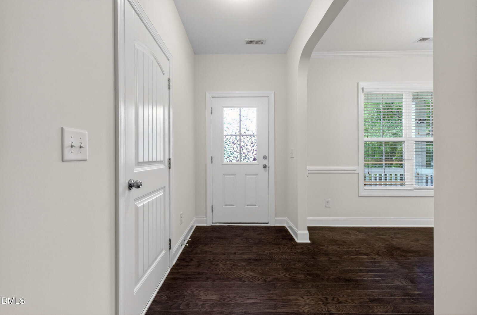 A bright entryway with dark wood floors, a white door with frosted glass, white walls, and a glimpse of a window with blinds in an adjacent room.