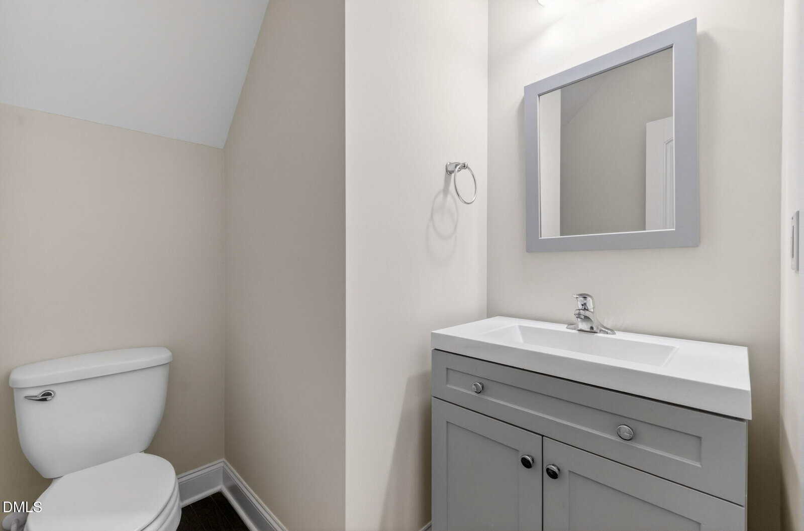 A small bathroom with a white toilet, a gray vanity with a white sink, a rectangular mirror above, and light-colored walls. The vanity has two drawers and cabinet doors. The bathroom has a sloped ceiling.
