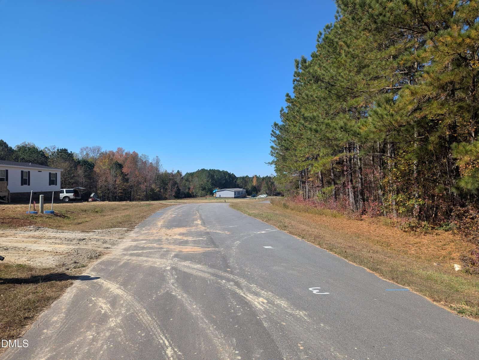 A paved road runs through a rural area with trees on the right and a few houses and cars on the left under a clear blue sky.
