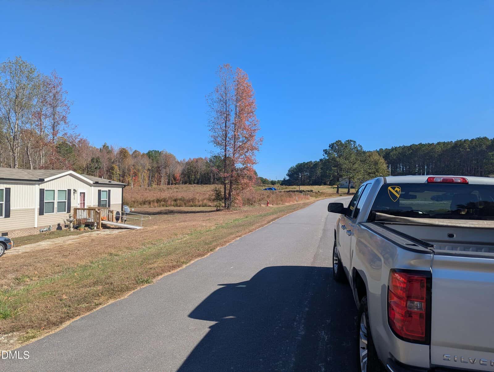 A white pickup truck is parked on a rural road beside a manufactured home, with fields, trees, and a clear blue sky in the background.