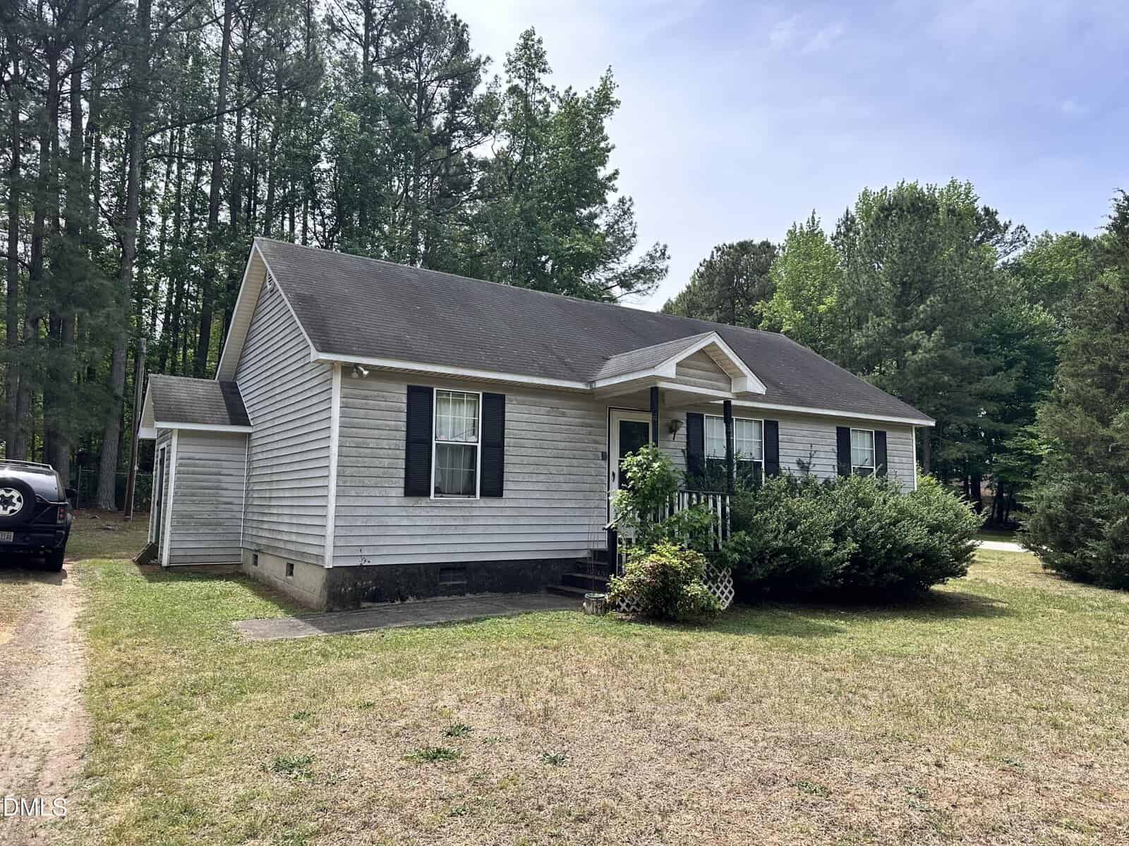 A single-story house with gray siding and black shutters sits on a patchy lawn, surrounded by trees. There are bushes near the front porch and a black vehicle parked to the left. The sky is partly cloudy.