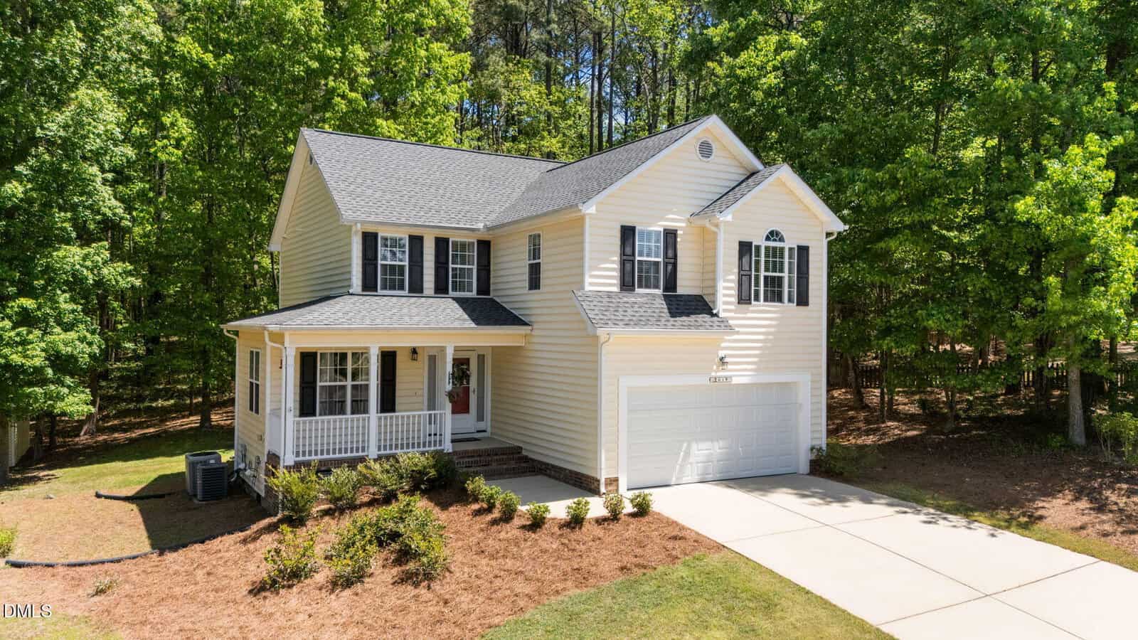 Two-story cream-colored house with dark gray roof, attached two-car garage, front porch, and driveway. The home is surrounded by green trees and has landscaped shrubs and mulch beds in front.