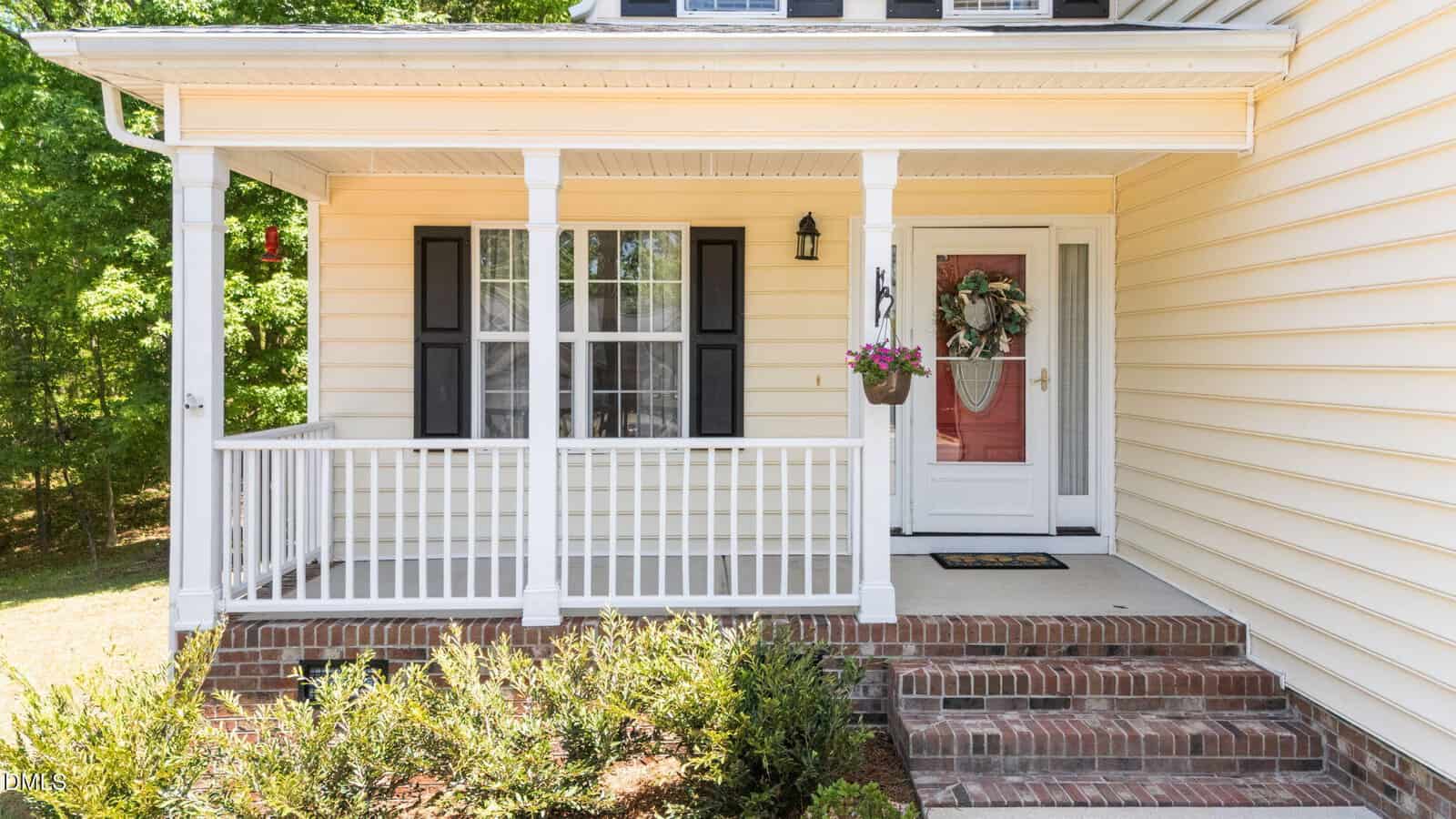 A yellow house with white trim, brick steps, and a covered front porch. The porch has white railings, a black lantern, a potted plant, and a front door decorated with a floral wreath. Shrubs line the walkway.